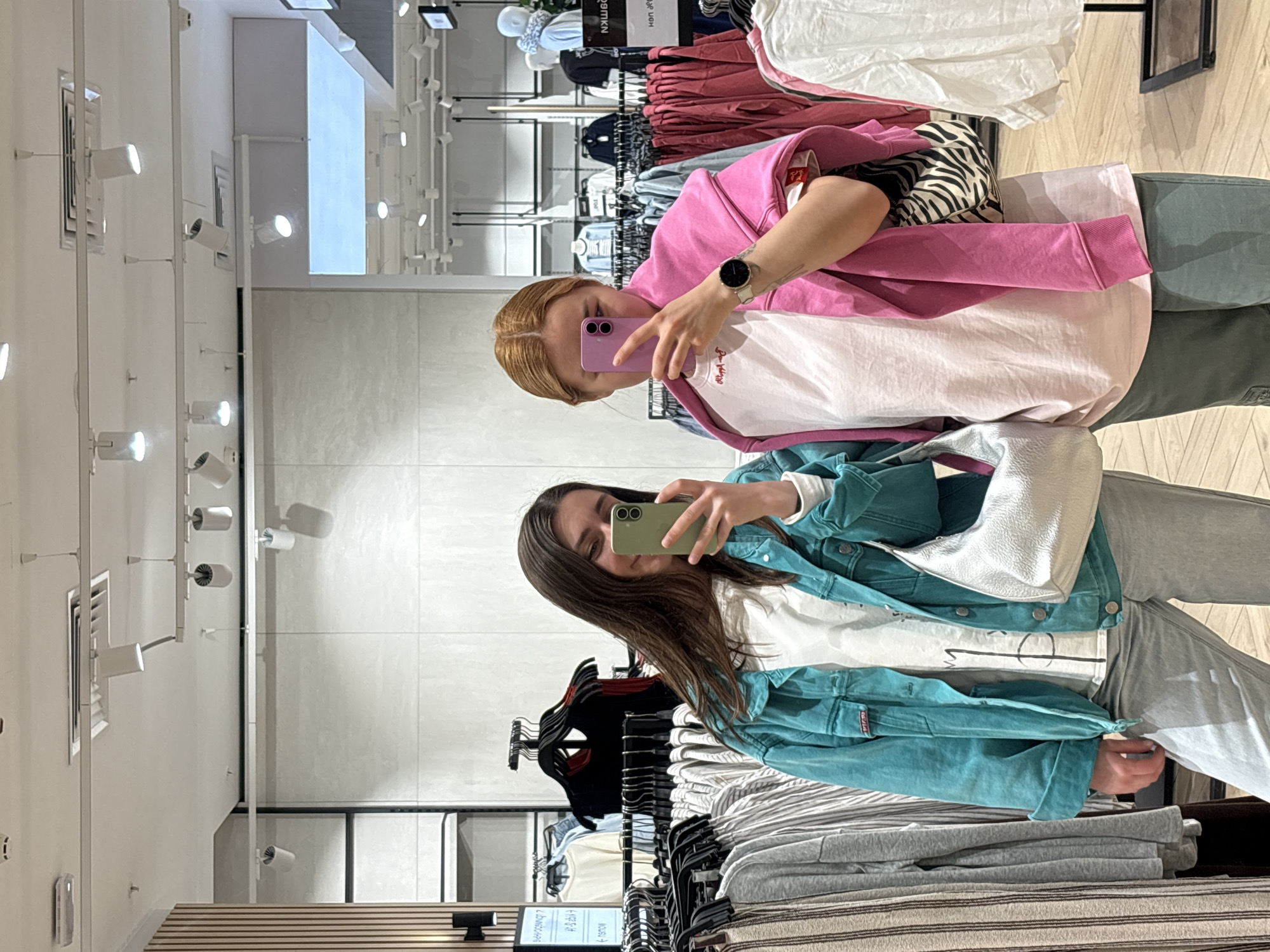 Two young women are taking selfies in what appears to be a clothing store. The woman on the left, with long brown hair, is wearing a blue denim jacket over a white t-shirt and grey sweatpants. She is holding a green smartphone. The woman on the right, with red hair, is wearing a pink sweatshirt over a white shirt and green pants, and is holding a purple smartphone. They are both looking at their phones. The store features racks of clothing in the background, including t-shirts, sweatshirts, and jackets. There are also visible lighting fixtures on the ceiling. The photo was likely taken in Minsk, Belarus.