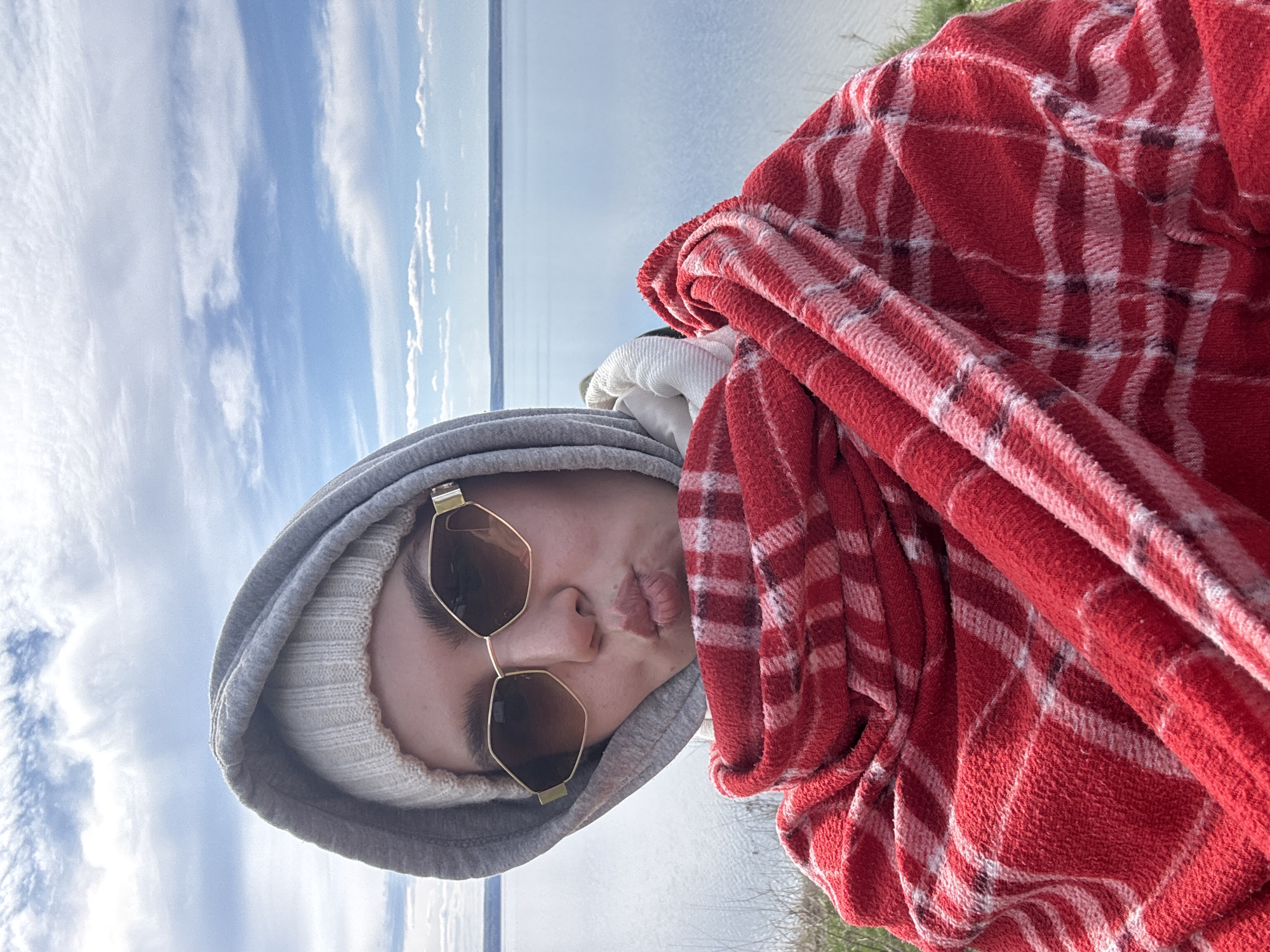 A selfie captures a person with fair skin, wearing a gray hoodie, a white knit cap, and sunglasses with gold frames. They are puckering their lips for the photo. A red and white plaid blanket is draped over their right shoulder. The background shows a body of water and a sky with clouds. The location is unknown, as the city is labeled Unknown, Unknown.