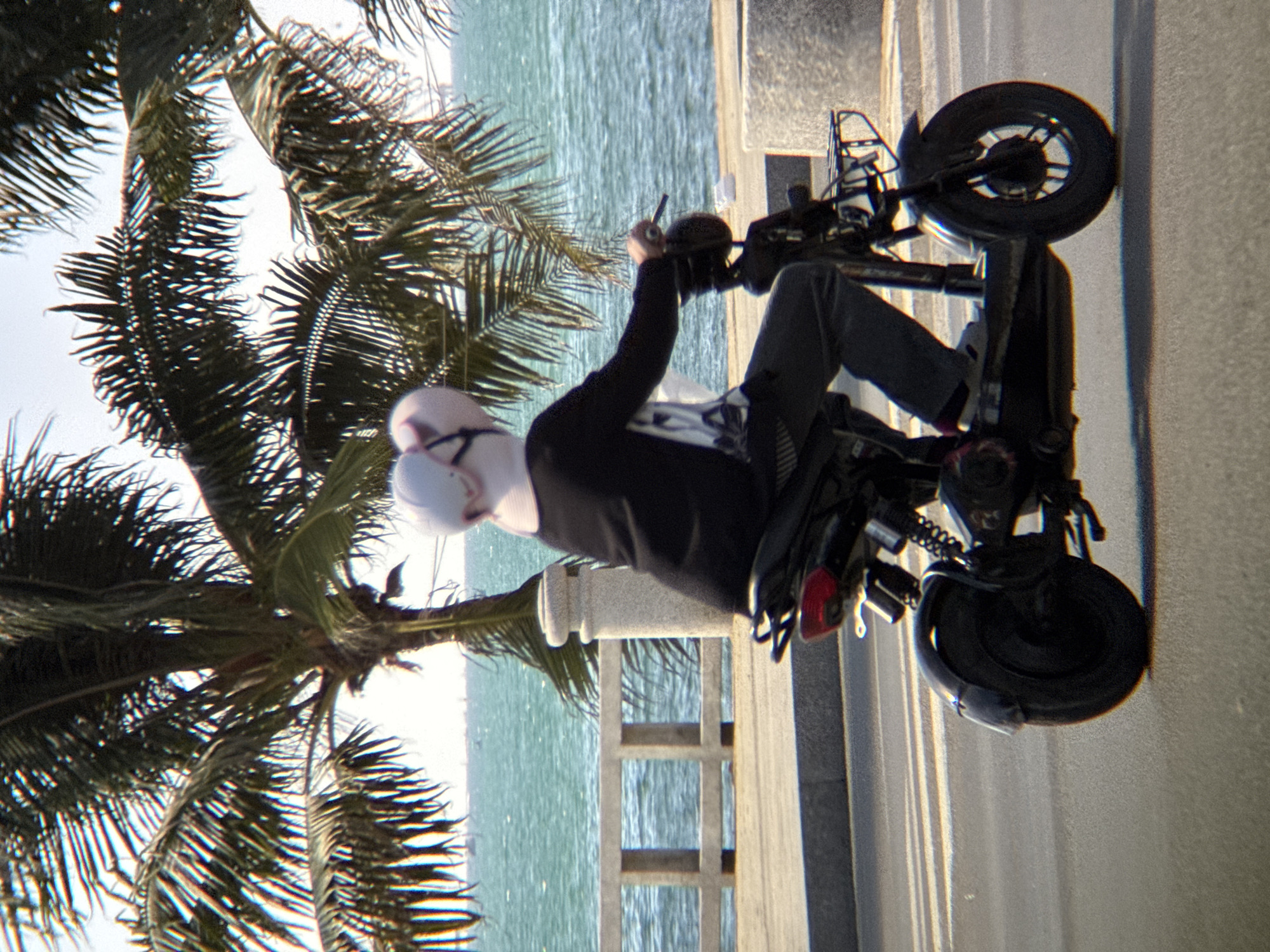 A person wearing a white and pink hat and a black jacket rides a black electric scooter on a paved road. The scooter has a black seat, a metal basket on the front, and a red taillight. In the background, palm trees with fronds partially obscure a turquoise ocean and a distant coastline. A concrete barrier with horizontal railings runs along the edge of the road, separating it from a sandy area and the sea. The scene is indicative of a location along the coast of Nha Trang, Vietnam, likely near the beach or promenade, given the presence of palm trees and the seaside environment. The lighting suggests it is daytime, with sunlight casting shadows.