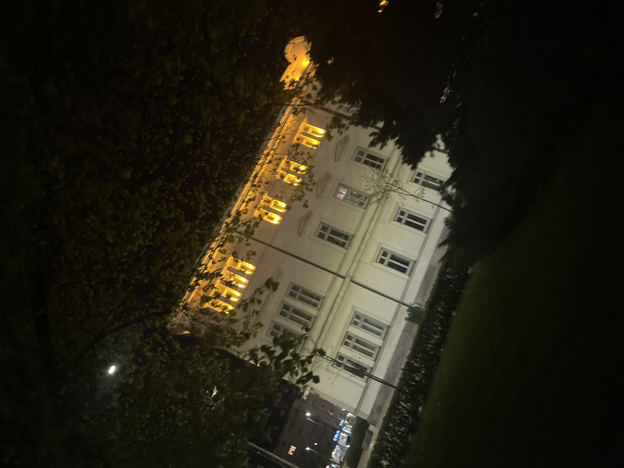 The image depicts a building at night. The building has multiple stories with windows illuminated by warm light. Tree foliage partially obscures the view of the building. In the lower part of the image, lights from other buildings and streetlights can be seen. The image appears to be taken from a low angle, looking up at the building. The location is Sofia, Bulgaria.