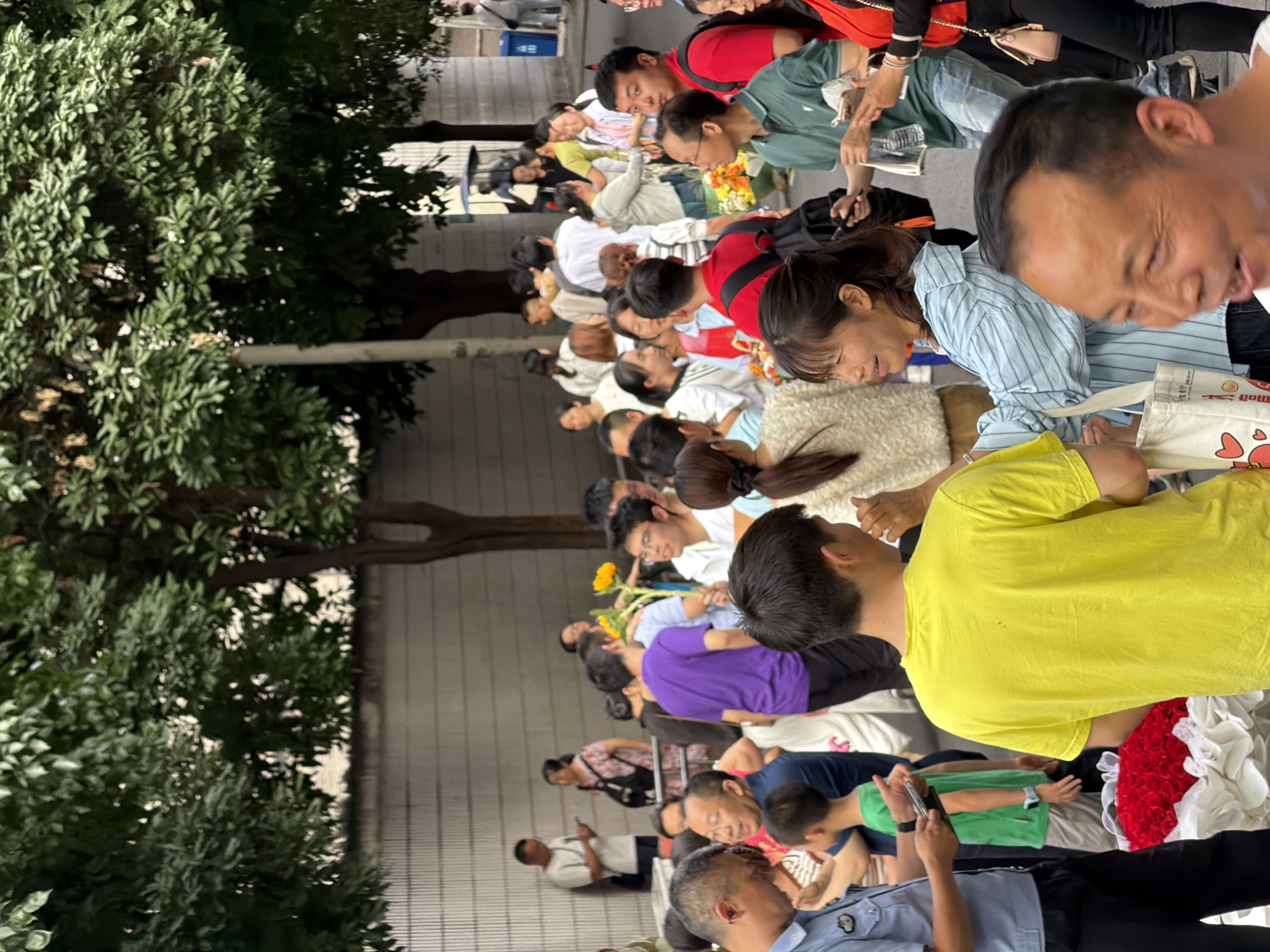 A dense crowd of people, predominantly East Asian, are gathered on a paved street in front of a concrete wall, possibly on the outskirts of a city. Several individuals are holding bouquets of flowers, notably a large arrangement of red roses at the forefront. Some people are looking at phones, while others are engaged in conversation. The presence of trees suggests an outdoor urban environment. The individuals appear to be of various ages and genders, dressed in casual clothing. The overall atmosphere seems to be one of a public gathering or event.
