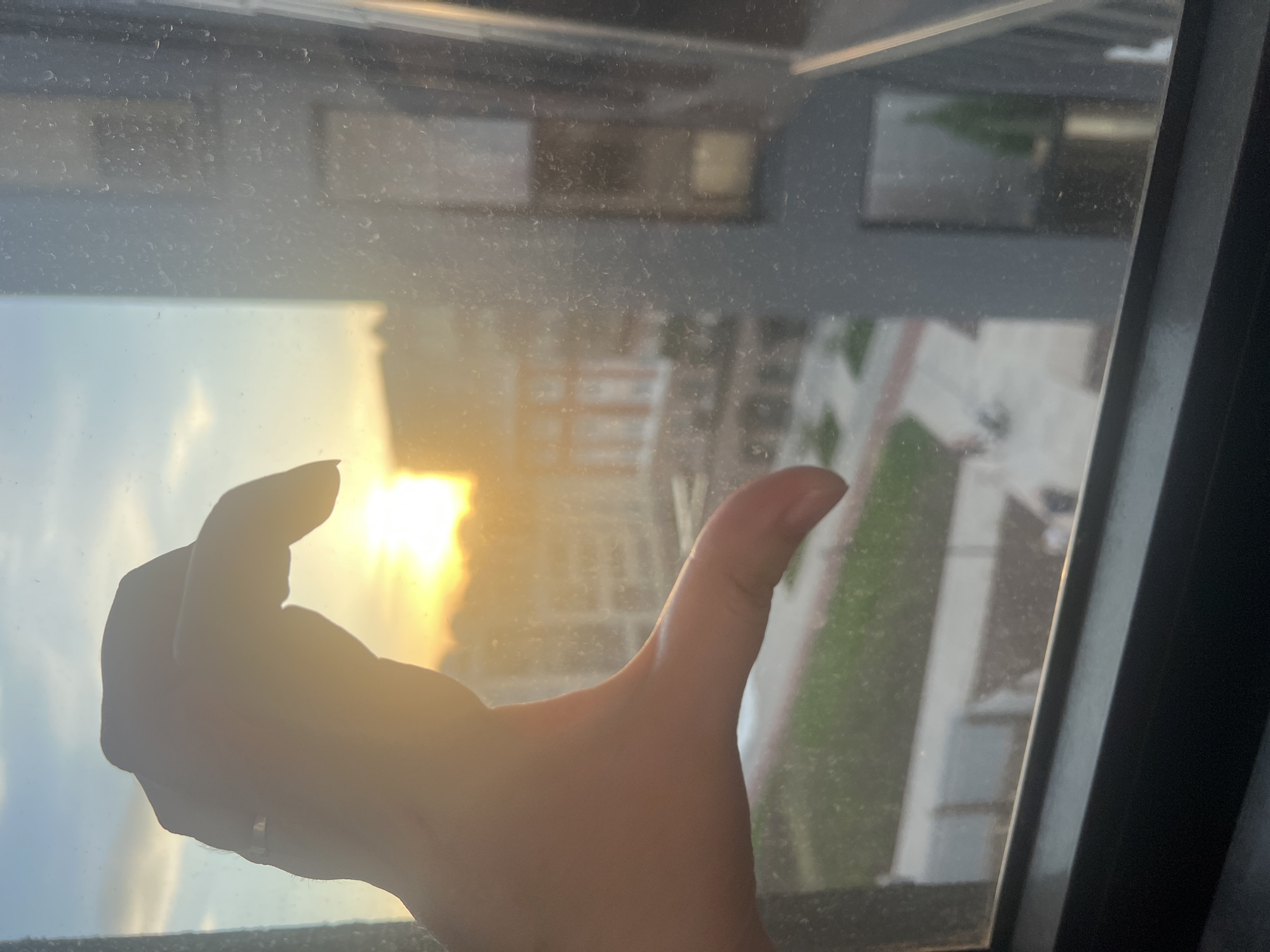 A hand is holding up a sun inside of a dirty window. The window appears to be facing a street with grass and buildings.
I am unable to determine the location is Izhevsk, Russia.