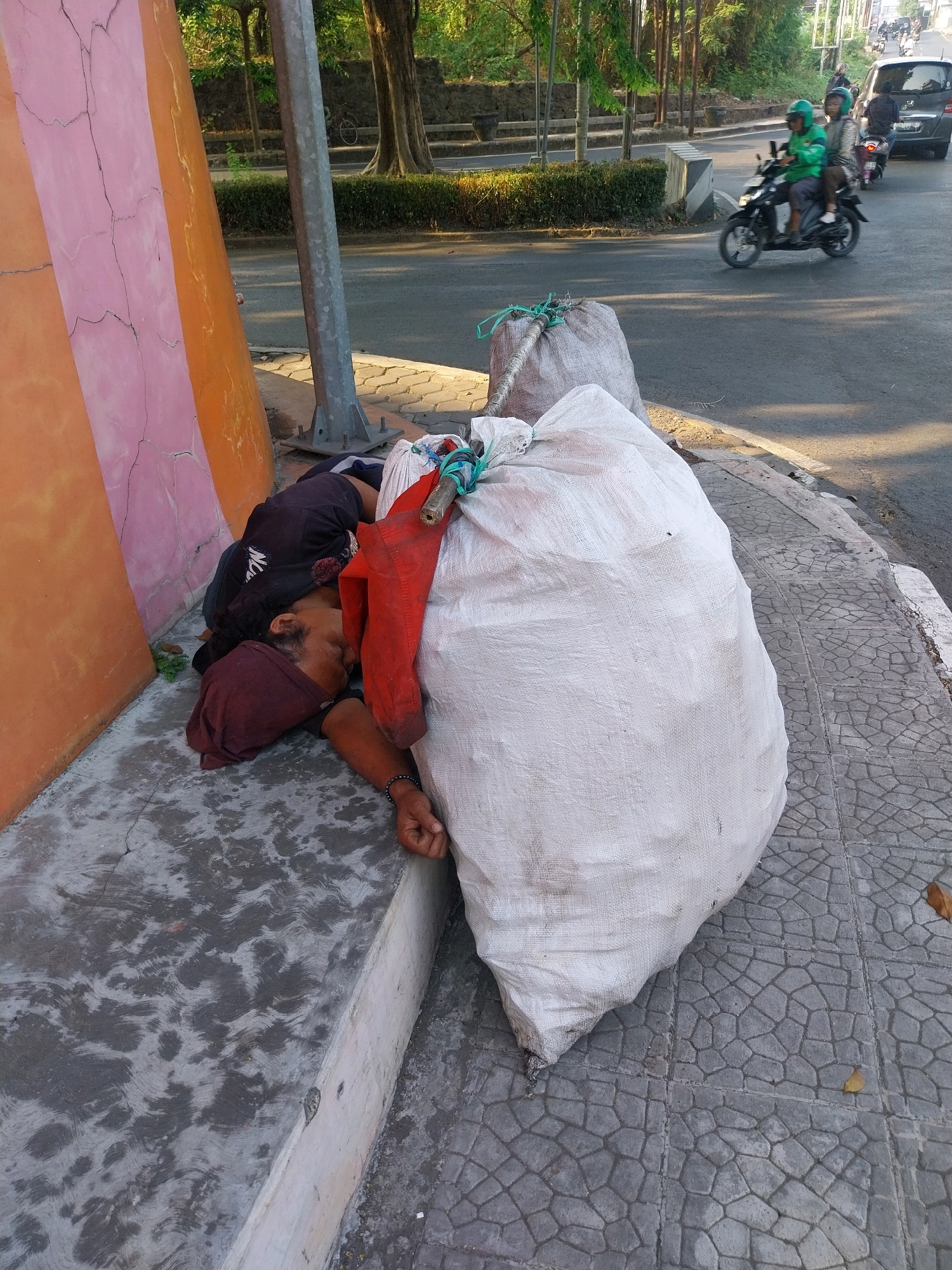 A man is sleeping on a concrete ledge next to a large white sack and a pink and orange striped pillar. He is wearing a dark shirt and a maroon headscarf. His arm is extended, resting near the sack. The scene appears to be outdoors, with a paved walkway and a road visible in the background. There are trees and buildings in the distance, suggesting an urban environment. Two people on a motorcycle are passing by on the road, and a dark-colored car is also visible. The lighting suggests it is daytime, with sunlight casting shadows. The overall impression is of a street scene in Cirebon, Indonesia, where a person is resting in a public space.