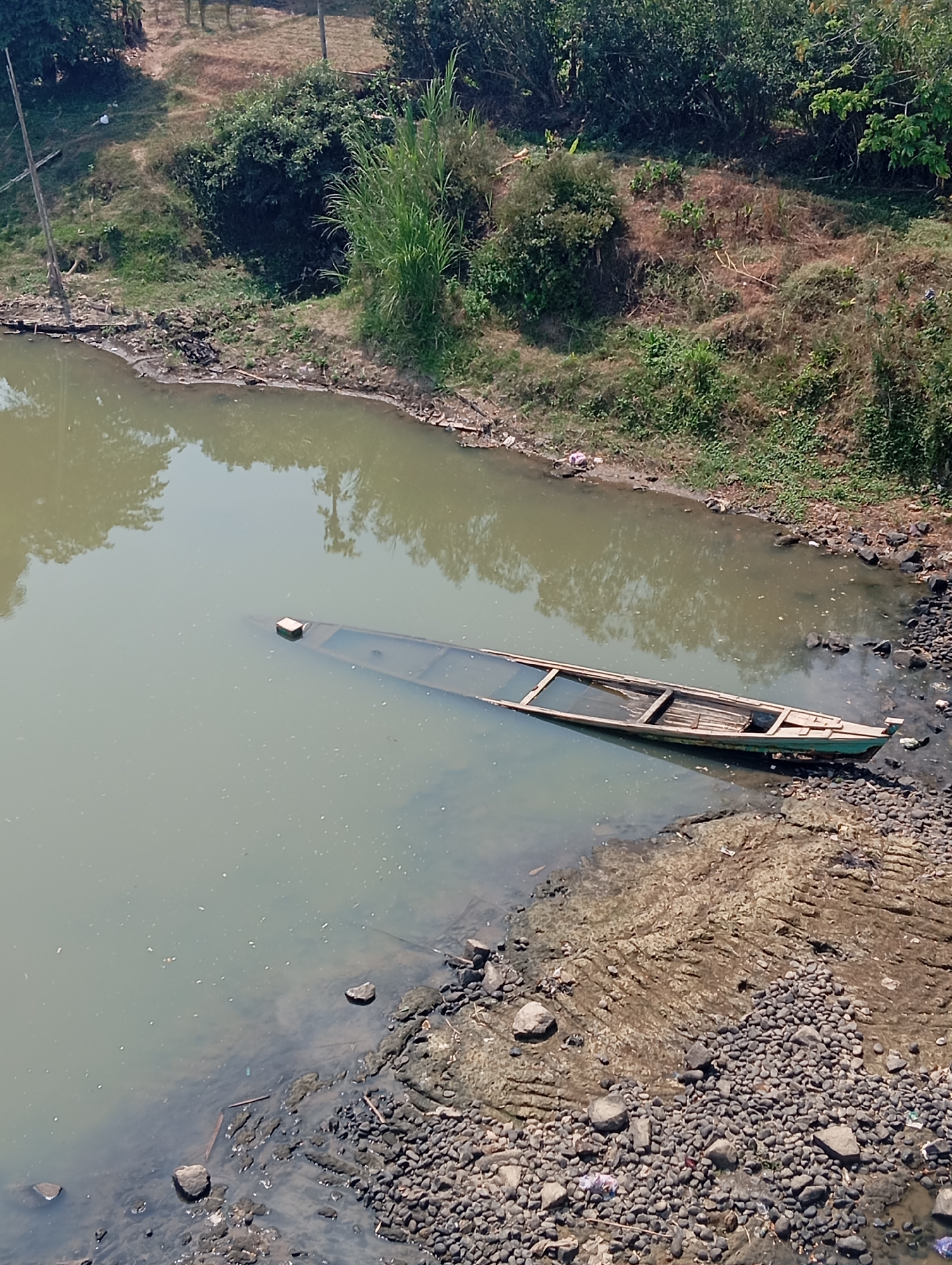In Bokol, Indonesia, a partially submerged wooden boat rests on the murky water's edge. The boat, painted green, is mostly underwater, with only its upper structure and a small square object visible above the waterline. The surrounding environment consists of a muddy bank strewn with rocks and debris, and lush green vegetation on the higher ground. The water is still, reflecting the surrounding trees and the clear sky.