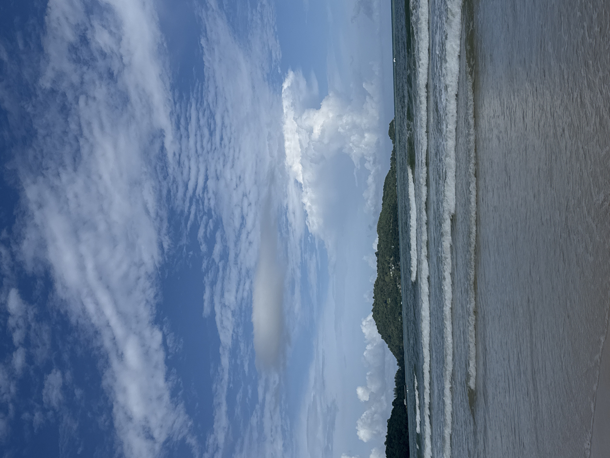 The image depicts a serene beach scene in Patong, Thailand, under a sky filled with dramatic clouds. The foreground is dominated by the gentle waves of the ocean lapping onto a sandy shore. The water is a deep blue-green, reflecting the sky and creating a sense of calm. Several distinct waves are visible, each with a white crest, suggesting a mild breeze.
In the mid-ground, two lush green islands rise from the sea. These islands are densely covered in trees, creating a verdant silhouette against the horizon. The larger island in the center features buildings nestled amongst the foliage, indicating a settled area. A smaller island is visible to the right of the main island.
The sky is a striking feature of the image, with a vibrant blue backdrop punctuated by a variety of clouds. Wispy cirrus clouds stretch across the upper portion, while larger, more dramatic cumulus clouds form a towering white mass in the center-right, hinting at potential weather activity. A single, elongated grey cloud floats higher in the sky towards the left.
There are no people or explicit actions visible in the frame, creating a peaceful and natural ambiance. The overall impression is one of tropical beauty and tranquility, characteristic of the beaches in Patong, Thailand.