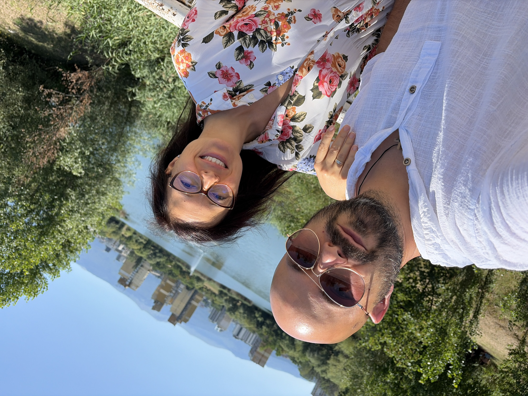 A couple is taking a selfie outdoors with a lake and buildings in the background, likely in Sofia, Bulgaria. The man in the foreground has a shaved head, beard, and is wearing sunglasses and a white collared shirt. The woman next to him is wearing glasses and a white dress with a floral print. Her right hand rests on his shoulder. The background shows a body of water with a fountain, trees, hills, and several multi-story buildings. The sky is clear and blue, suggesting a sunny day.