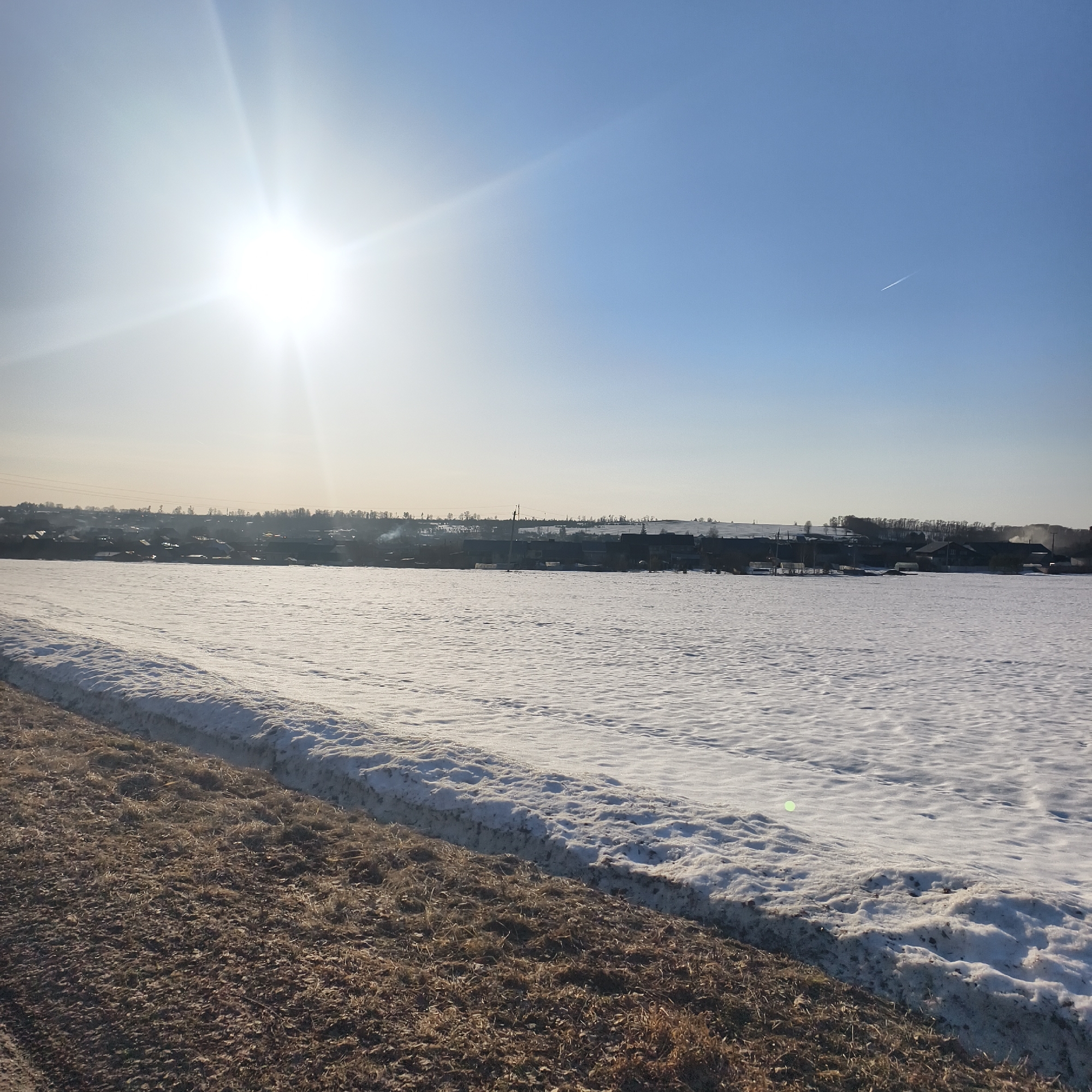 A daytime shot depicts a rural landscape under a bright sun and clear blue sky in Фоки, Russia. A snow-covered field dominates the foreground, with a strip of brown, possibly dirt or dry grass, running along the lower left. In the distance, a line of buildings, possibly a village or small town, is visible against the horizon. The sun is high in the sky, with its glare creating a bright, almost washed-out effect in the upper portion of the image. A contrail from an airplane can be seen in the upper right quadrant of the image. The overall impression is one of a cold, sunny winter day in a sparsely populated area.