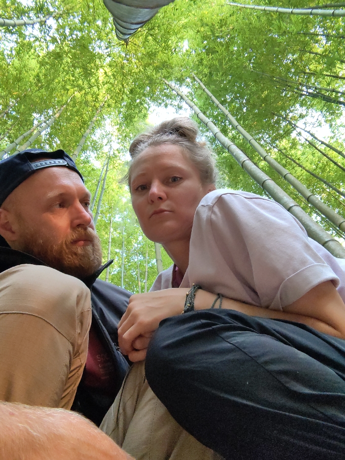 A low-angle photograph captures two individuals within a dense bamboo forest in Mù Cang Chải, Vietnam. On the left, an adult male with a reddish-brown beard and a backward dark baseball cap wears a dark jacket and khaki pants; he looks towards his left, away from the camera. On the right, an adult female with blonde hair pulled into a bun wears a light-colored short-sleeved top and dark pants; she gazes directly into the camera with a neutral expression. Her left hand rests on the man's leg, and she wears a dark braided bracelet with beads on her left wrist. The background consists of numerous tall, slender green bamboo stalks and lush foliage, ascending towards a bright, overhead sky. A dark, indistinct object is partially visible at the top center of the frame.
