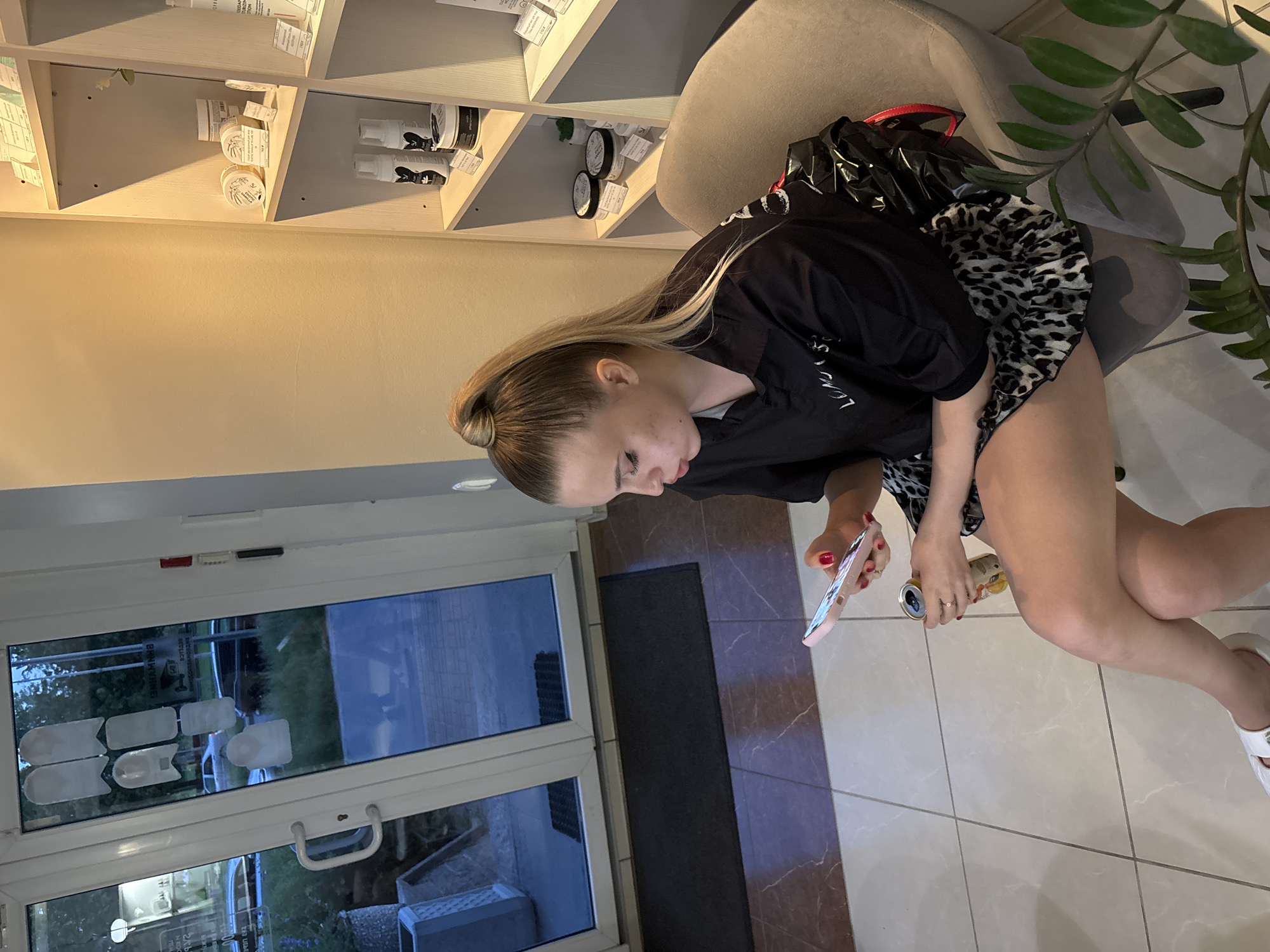 In Romanovka, Russia, an indoor scene depicts a young woman seated on a light grey upholstered chair, positioned to the right of a large white-framed glass double door. The woman, with blonde hair tied in a high ponytail, wears a black collared shirt with partially visible white lettering ("LUMOUS") on the left chest and a leopard print ruffled skirt. She has red nail polish on her fingers and white open-toed sandals on her feet, with her legs crossed at the knees. Her gaze is directed downwards at a pink smartphone held in her left hand, while her right hand holds a golden-yellow beverage can. A black plastic bag is draped over the back of her chair, partially obscured.
The interior features a tiled floor composed of light grey/white sections in the foreground and darker reddish-brown sections with lighter veins closer to the door. The wall to the left is plain beige, while the wall to the right features light-colored shelving units displaying various cosmetic or personal care products in white and black containers, some with visible price tags. A small green plant is situated on the lower shelf, and a larger green leafy plant (possibly a ZZ plant) occupies the lower right foreground.
Through the glass double door, the exterior is visible, showing a dark paved area, green grass, and trees under what appears to be evening or twilight lighting. A silver sedan and a dark-colored SUV are discernible in the distance. Reversed text, including "ВАНИЛА" and "BRANIMANHA", is visible on the glass due to reflections or interior signage. Several opaque white, irregularly shaped stickers are affixed to the glass panels. A dark grey doormat rests immediately inside the door.