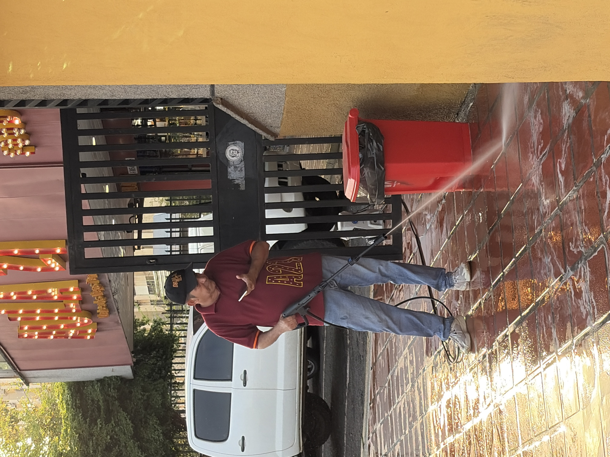 This image, set in Guayana City, Venezuela, depicts a man actively engaged in pressure washing a tiled ground surface. The scene appears to be an outdoor area, possibly a business entrance or patio, bordered by a black metal gate and a yellow building wall.
The central subject is a man, likely middle-aged, wearing a maroon short-sleeved polo shirt with a gold "AZ" or "A7" design on the front, blue jeans, and light-colored sneakers. He also wears a dark baseball cap. In his right hand, he holds a black and blue pressure washer gun, directing a stream of water onto the reddish-brown tiled floor, which is visibly wet and reflective. A white cigarette is held between the fingers of his left hand.
Behind the man, a white pickup truck is partially visible. To his right, behind the gate, stands a bright red trash can with a black garbage bag liner. On the far left, a sign adorned with yellow and red exposed light bulbs is visible, and some indistinct gold text can be faintly seen on it, possibly reading "FRESH" or a similar word, along with what might be "100%." The ground is a grid of square, reddish-brown tiles, glistening from the water. Green foliage and a building with a pink wall are seen in the background on the left.
The lighting suggests it is daytime, with sunlight reflecting off the wet surfaces. The man appears focused on his task. A small circular sticker with an unclear logo or face is affixed to the black gate. The activity indicates maintenance or cleaning work is underway.