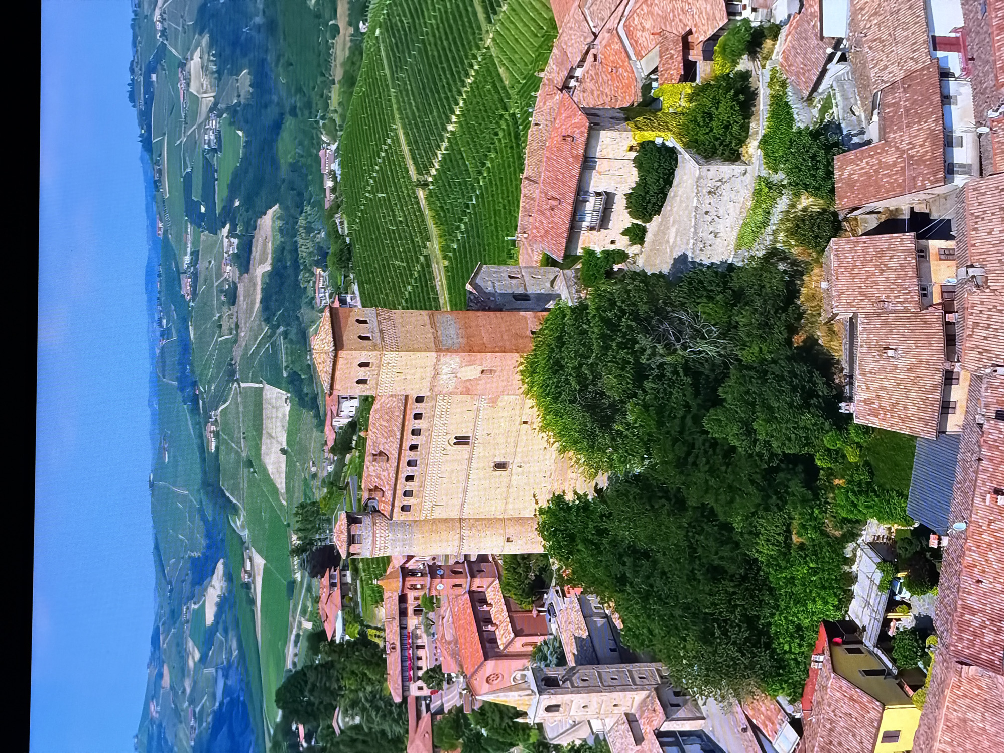 The image is an aerial view of a historic Italian village nestled within rolling hills covered in vineyards. The village features a prominent brick castle or fortified tower as its central element, surrounded by terracotta-roofed houses and a church with a tall, slender bell tower. Lush green trees and dense vegetation are interspersed throughout the village and on the surrounding hillsides. The landscape exhibits a well-maintained agricultural pattern, with neat rows of grapevines dominating the background, suggesting a wine-producing region. The sky is a clear, bright blue. The scene is tranquil and picturesque, depicting a preserved historical settlement in a scenic rural environment. The requested location in Chirchik, Uzbekistan, is not present in this image.
