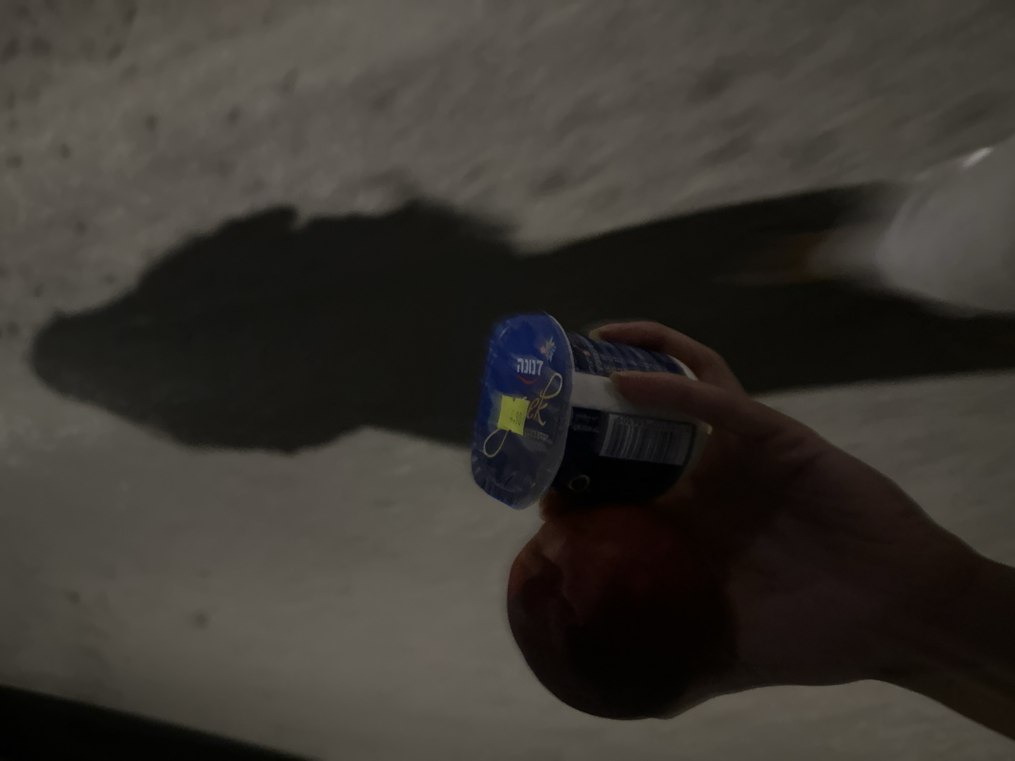 In the city of Tel-Aviv, Israel, a hand is holding a peach and a small cup of yogurt. The yogurt cup has a blue lid with a yellow price tag. The background is a blurry, speckled surface, possibly a sidewalk or wall, with a long shadow of a person cast across it. The lighting suggests it is evening or nighttime.