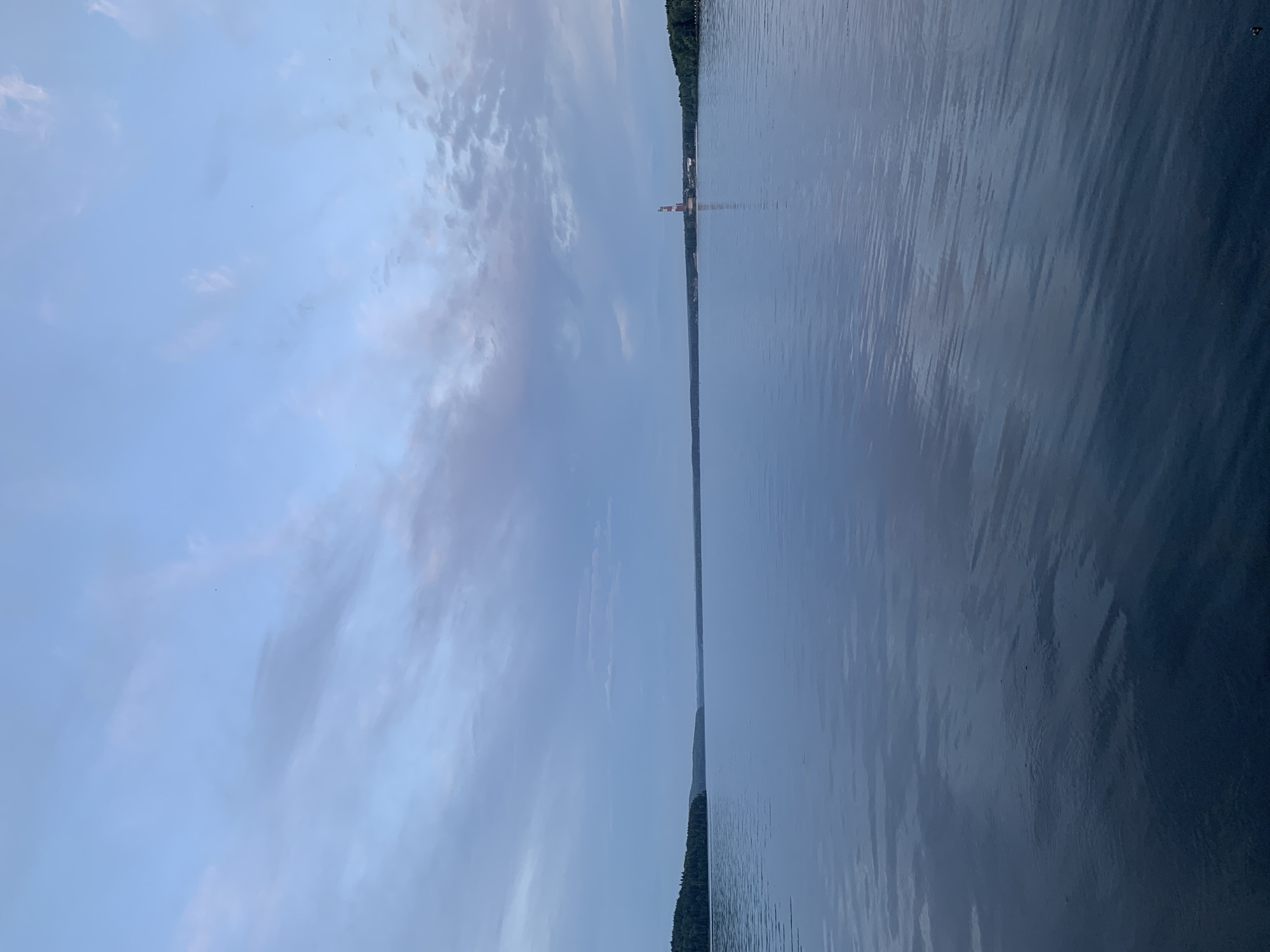 This is a landscape photograph taken from a body of water, likely a river, looking towards a distant shoreline. The image is oriented vertically, with the sky occupying the left half and the water filling the right half.
The sky is a pale blue with scattered wispy clouds, suggesting it is either early morning or late afternoon. The water is calm and reflects the sky, creating a smooth, dark surface with subtle ripples. The distant shoreline is lined with dark green trees, indicating a natural or semi-rural setting. A prominent feature on the distant shore is a tall, red and white structure, possibly a lighthouse or a radio tower, which stands out against the tree line. There are no visible people in the photograph. The overall mood is peaceful and serene. The location is identified as Chaikovsky, Russia.