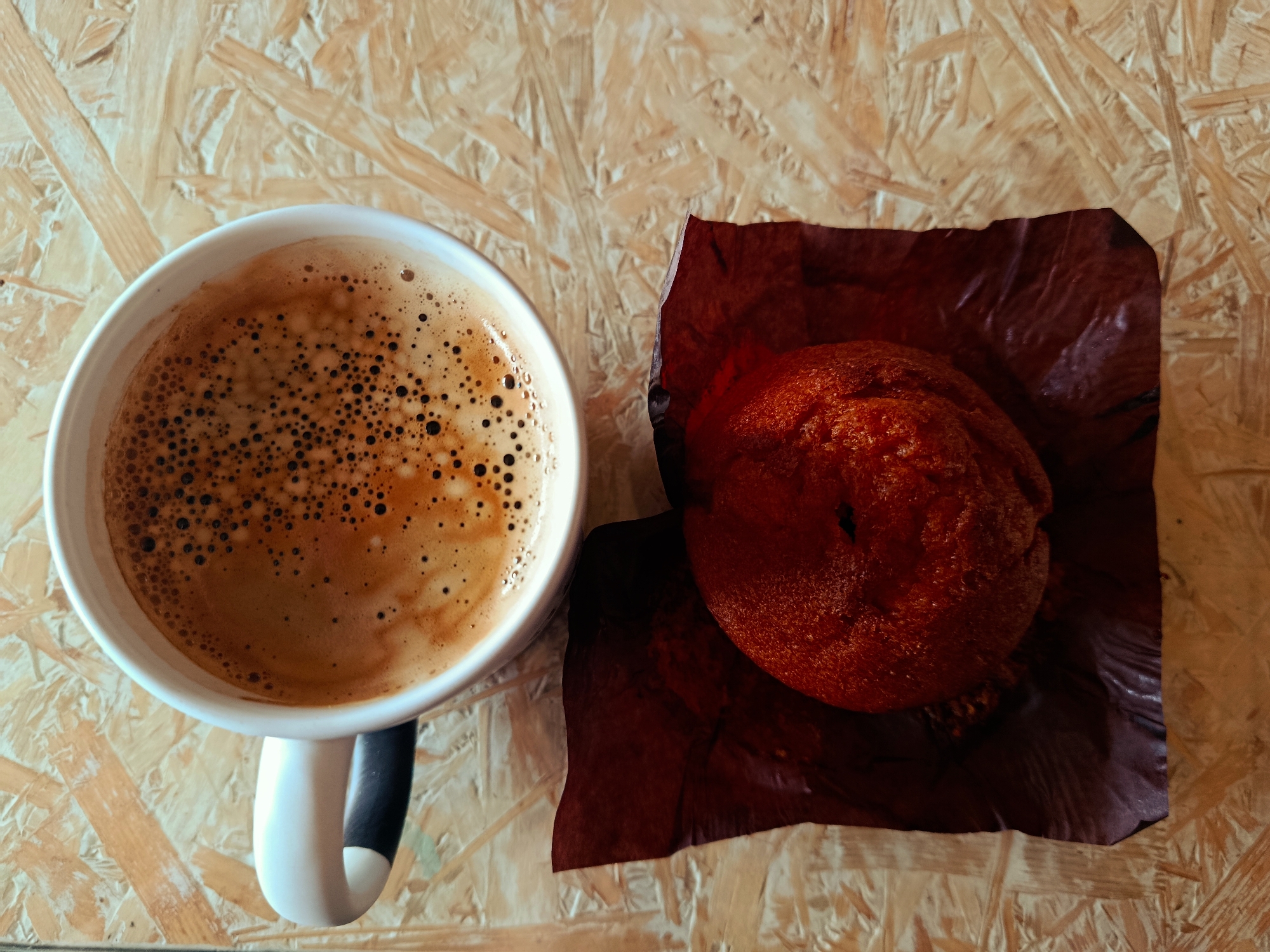 An overhead shot shows a white coffee mug filled with dark coffee with a foamy crema, positioned to the left of a reddish-brown pastry wrapped in dark red paper. The mug and pastry rest on a light brown, textured surface composed of wood chips.