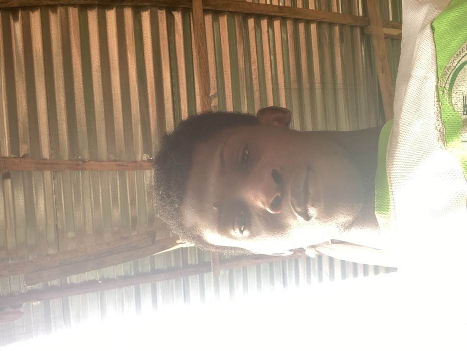 The image displays a young adult male located in Agege, Nigeria. The subject has dark skin, short, dark, curly hair, and faint stubble on his chin and jawline. He is wearing a white shirt, possibly mesh fabric, with green trim visible on the collar and upper chest, and a partially discernible green logo. The subject faces directly towards the camera with a neutral expression. A bright light source is positioned to the left, causing significant overexposure on the left side of the subject's face and upper body. The background features a corrugated metal roof structure with horizontal ridges, supported by rough-hewn wooden rafters positioned diagonally and horizontally across the upper portion of the frame.
