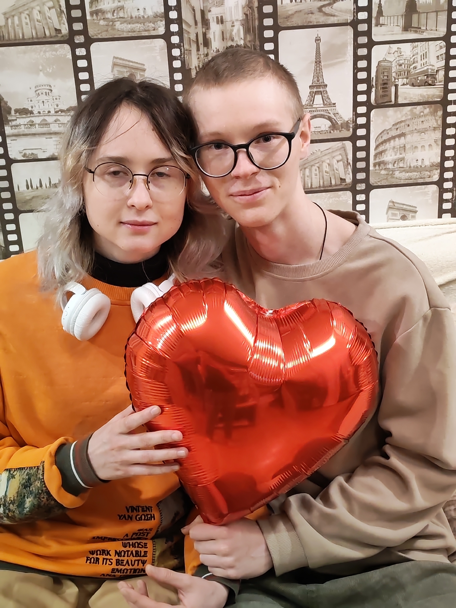Two individuals, a man and a woman, are positioned together in what appears to be an indoor setting. Both are holding a large red heart-shaped balloon. The woman is on the left, wearing an orange sweatshirt with printed text on it, a black turtleneck underneath, and round glasses. White headphones are around her neck. The man, on the right, is wearing a beige sweatshirt and round glasses. In the background is a wallpaper depicting various landmarks. The location is Chaikovsky, Russia.