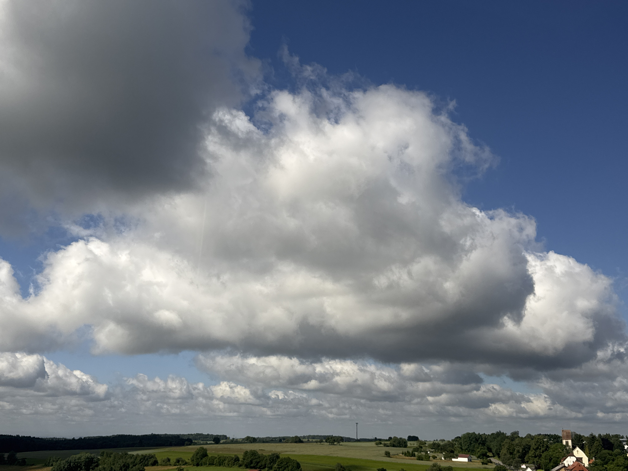 A wide-angle landscape photograph captures a dramatic sky over the town of Strümpfelbrunn, Germany. The sky is dominated by a large, textured cloud formation that spans across the upper portion of the image, with varying shades of gray and white suggesting depth and density. Smaller, scattered clouds are visible below this main formation, with patches of clear blue sky interspersed. The lower third of the image reveals a rural landscape. A tree line borders the horizon, with a few scattered buildings visible, including a white church with a red-brown roof and steeple, and other red-roofed houses. Fields and rolling green hills are visible in the foreground, with scattered trees and a tall, thin structure, possibly a radio tower, in the distance. The overall impression is of a bright, overcast day in a German countryside.