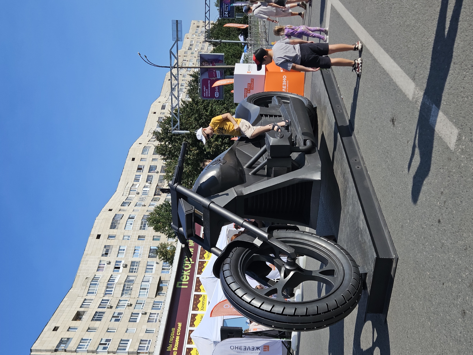 On a sunny day in Ulyanovsk, Russia, a large, black, non-functional motorcycle sculpture is displayed outdoors on a street. The sculpture is the central focus, positioned on a black platform. A young boy in a yellow t-shirt and beige shorts is seated on the motorcycle, looking towards the right. To the right of the sculpture, a young boy in a grey t-shirt and black shorts stands facing away from the camera, looking at the motorcycle. Further to the right, a young girl in a purple outfit walks away from the camera. In the background, several other people are visible, along with tents and banners associated with an event. A multi-story residential building dominates the skyline, with trees and streetlights interspersed. The ground is asphalt, marked with white lines.