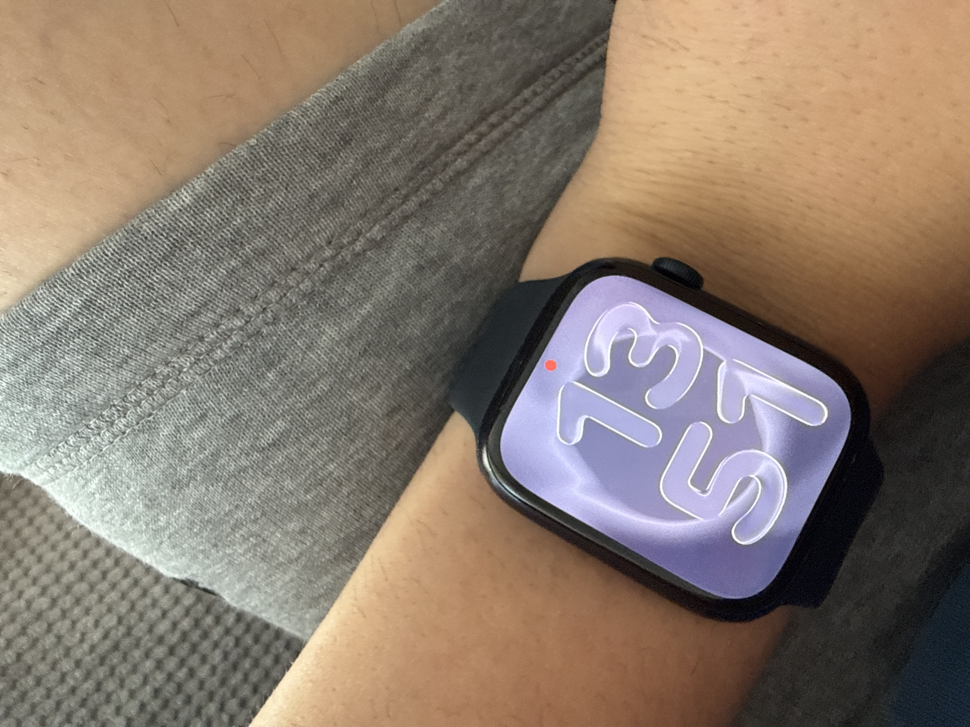 A close-up shot shows a person's arm wearing a black Apple Watch. The watch face displays the time as "13:53" in large, stylized white numerals on a light purple background. A small red dot is visible in the upper left corner of the watch face. The arm is positioned against a backdrop of gray fabric, likely clothing. The scene appears to be indoors and, without more context, there is no way to determine its location in the city of Lovech, Bulgaria, or to know whether it is in Lovech at all.