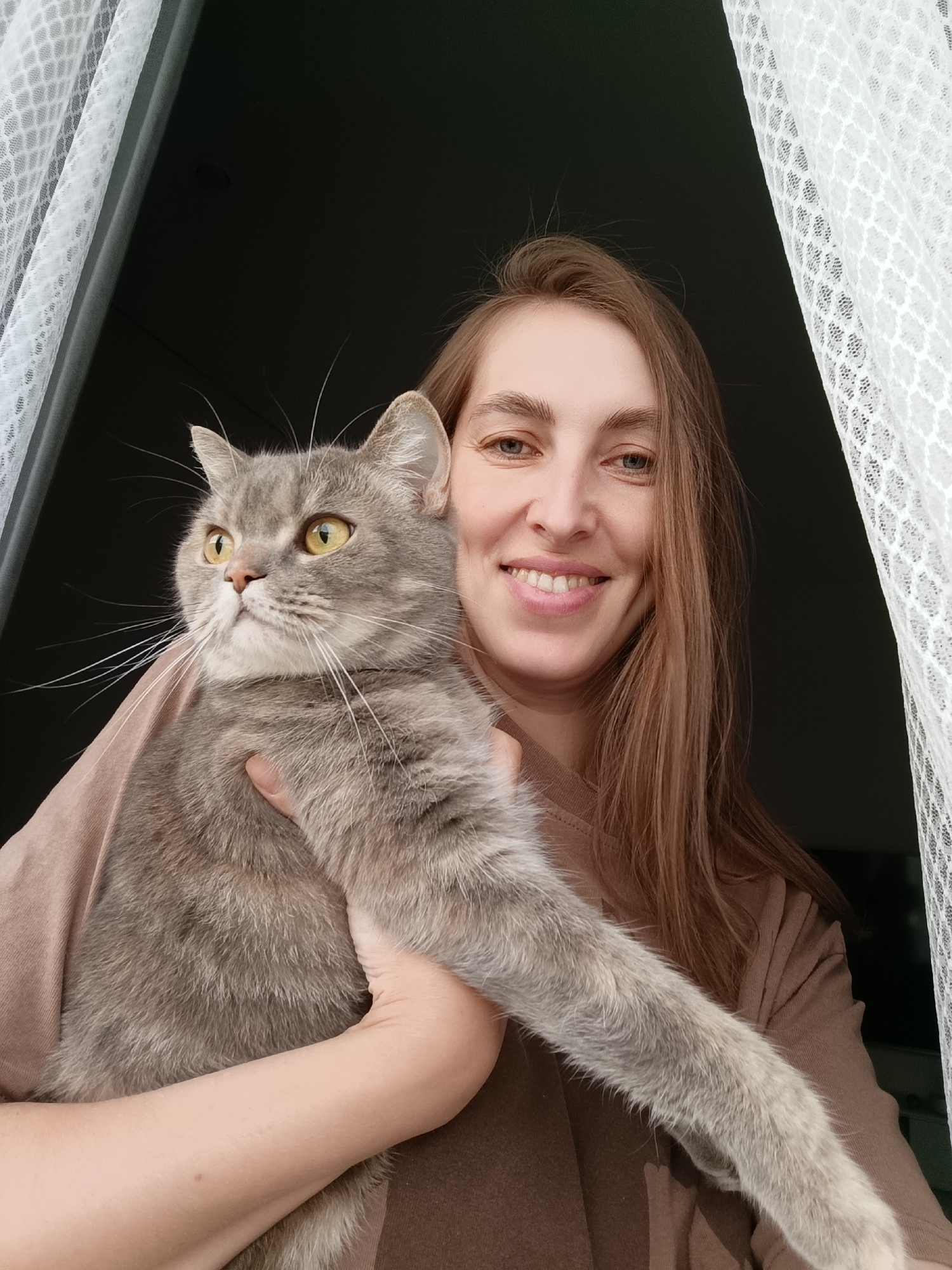 A young woman is holding a grey tabby cat. She is smiling and looking at the camera. The cat is looking to the left. They are both standing in front of a window with sheer white curtains. The background is dark. The woman has long, brown hair and is wearing a brown shirt. The cat has yellow eyes and a white chest.