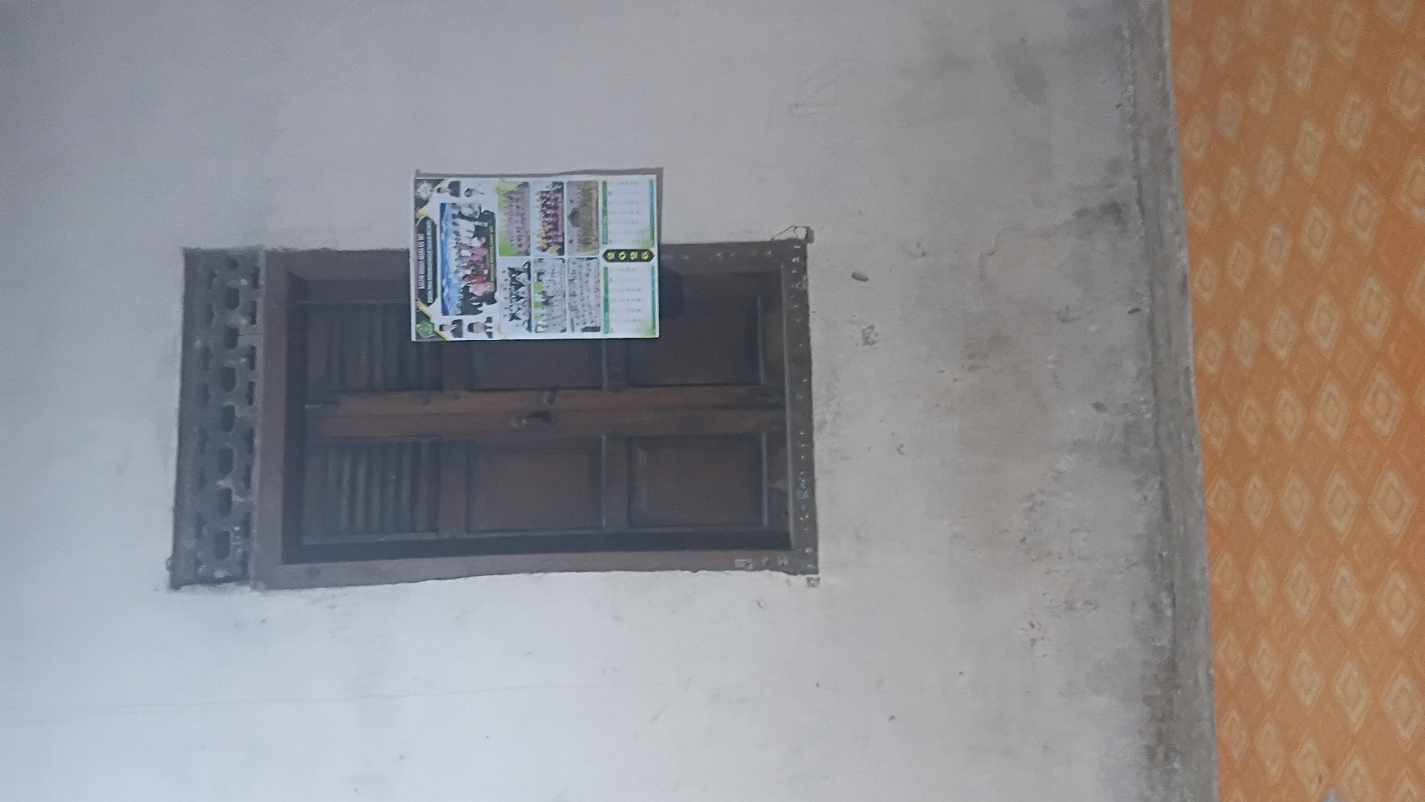 The image displays an interior scene featuring a wooden window with shutters, partially open. A calendar with multiple photographs is affixed to the wall to the right of the window. The calendar appears to be from the year 2023. The wall is a light, neutral color and shows some signs of age and wear. Below the wall, an orange tiled floor with a diamond pattern is visible. The lighting in the image is somewhat dim, suggesting it might be indoors or taken in low light conditions. The specific location of Prabumulih, Indonesia, cannot be determined from the visual information present in the image itself.