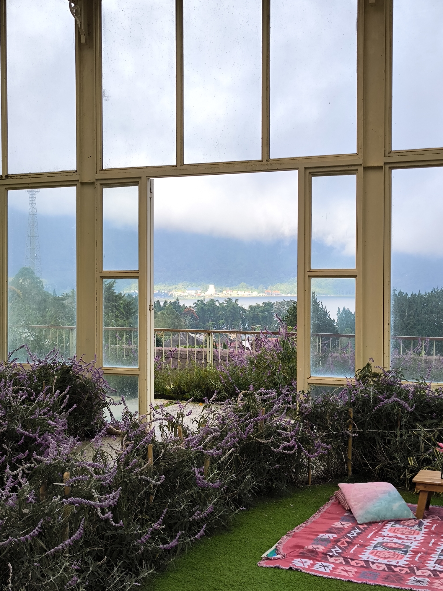 The scene depicts an indoor setting in Candikuning, Indonesia, looking out through a large window toward a lake and mountains on a cloudy day. The window, framed in light brown wood, consists of multiple panes, with the central section forming an open doorway leading onto a balcony with a metal railing.
Inside, a patch of artificial green grass hosts a pink patterned blanket, a turquoise gradient pillow, and a small wooden side table. Purple flowering plants, possibly lavender, fill the space immediately inside the window, creating a foreground border.
Beyond the balcony, the view encompasses a body of water, presumably a lake, surrounded by lush greenery and mountain slopes. In the distance, structures resembling buildings are visible, potentially part of a town or village along the shore. A tall communications tower stands on a hill in the left portion of the scene. The sky is overcast, and the light suggests a cool, possibly damp atmosphere. There are no people present in the photograph.