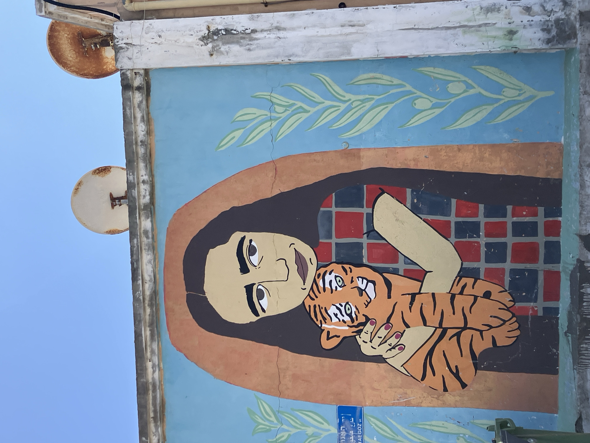 A mural in Tel Aviv, Israel, depicts a cartoonish person with dark hair and tan skin holding a tiger cub. The person is wearing a checkered shirt with red and black squares. The background is a light blue with green olive branches at the top. The mural appears on the side of a building with a rusted satellite dish attached. A street sign in Hebrew and English (Haegoz Street) is visible at the bottom.