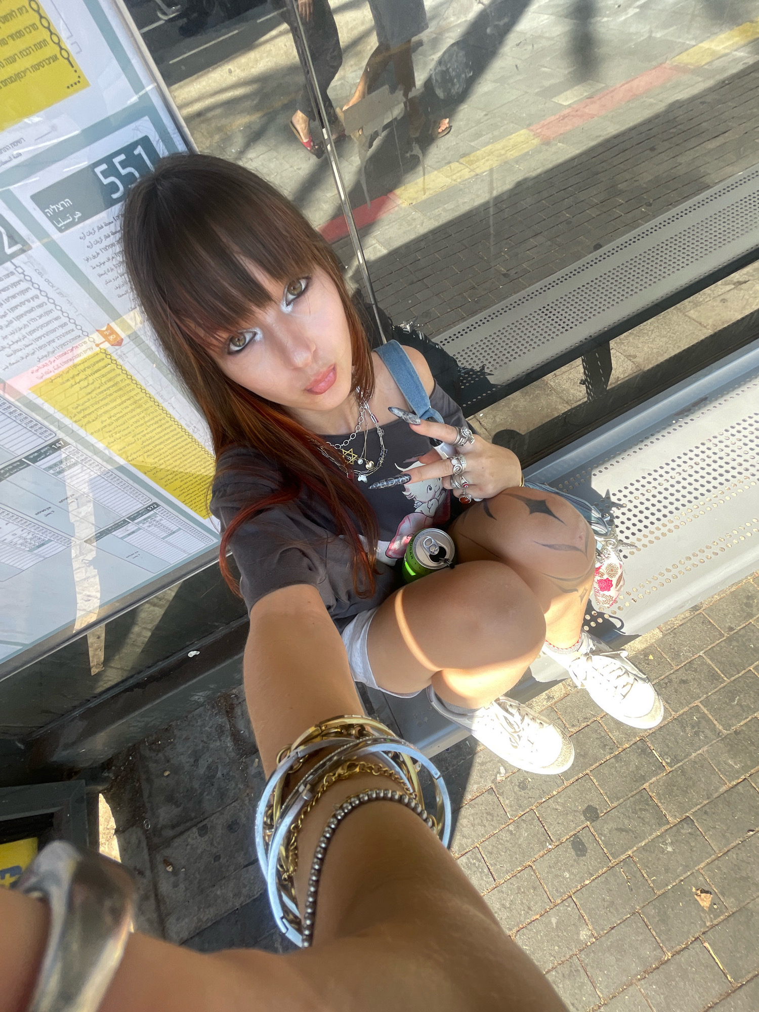 This image depicts a young woman sitting on a metal bench at a bus stop in Raanana, Israel. She is looking at the camera, with her tongue sticking out slightly. She has long brown hair with reddish highlights, and is wearing a dark grey t-shirt, denim overalls, and white sneakers. She has several rings on her fingers and a collection of bracelets on her wrist. There is a can of soda on the bench next to her, and a key chain with a pink and white design attached to her shorts. In the background, a bus schedule is visible, with route number 551 prominently displayed. The ground is paved with grey bricks. The lighting suggests it is a sunny day, with shadows cast by the bus stop structure and passersby.