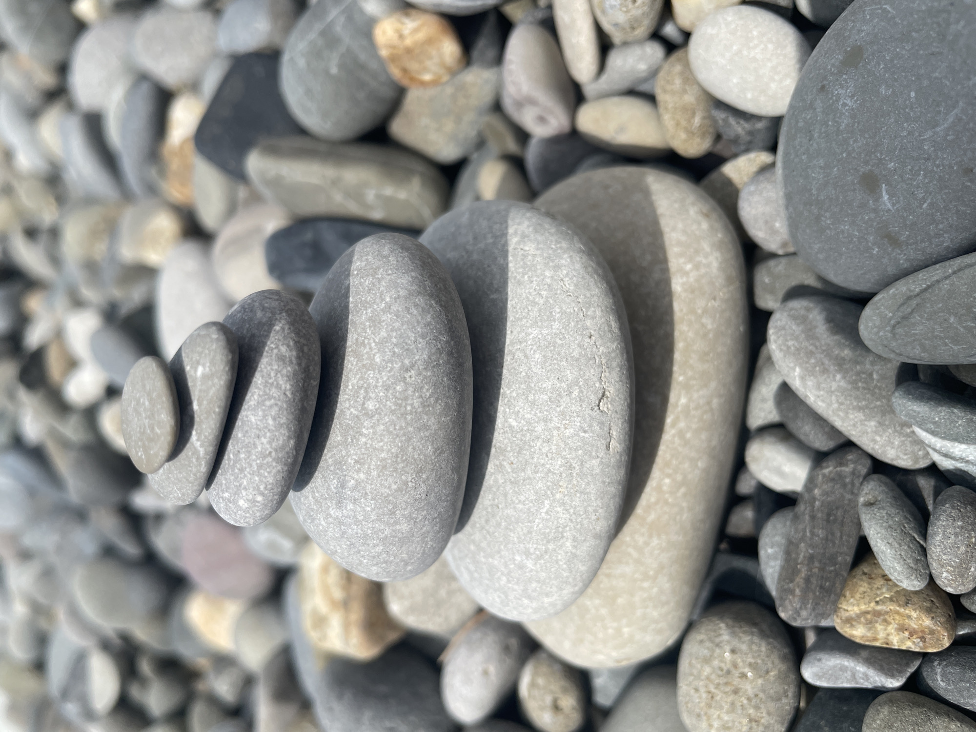 A close-up, eye-level shot captures a stack of five smooth, rounded pebbles on a beach. The pebbles are various shades of gray, with the largest, a pale tan, at the bottom, topped by four progressively smaller gray stones. The stacked pebbles are the main focus, appearing slightly blurred due to shallow depth of field. They rest on a bed of similar pebbles that fill the foreground and extend into the blurred background, suggesting a beach or rocky shoreline. The lighting appears to be natural daylight, casting subtle shadows. The image does not depict any people, actions, or interactions. The location is described as Unknown, Unknown.