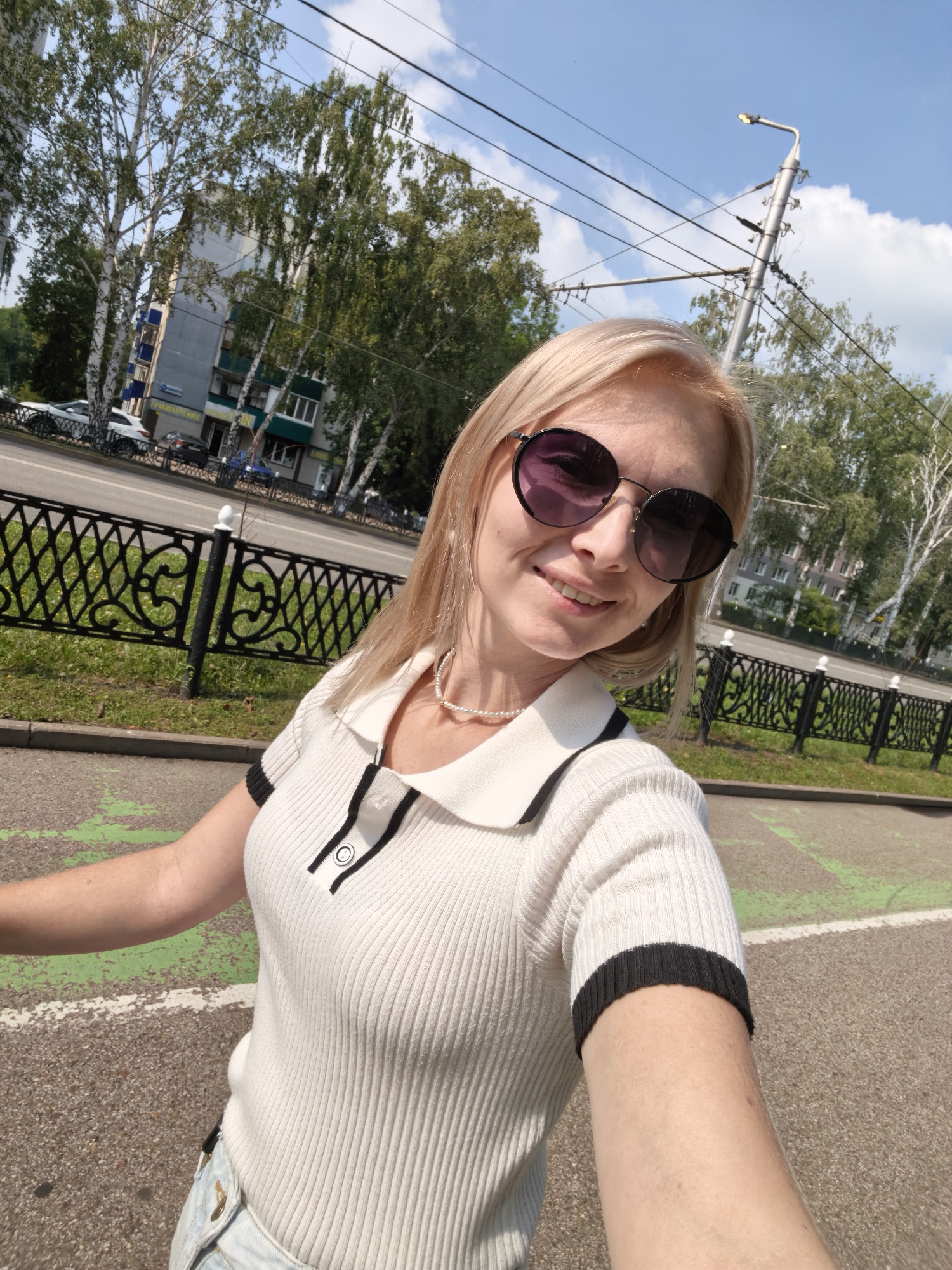 The image is a selfie taken outdoors, possibly on a sidewalk or path in the city of Sterlitamak, Russia. A woman with blonde hair and sunglasses is visible, wearing a white polo shirt with black trim. She is smiling and looking at the camera. In the background, there are trees, a street with cars, a building, a black metal fence, and a lamppost with overhead wires. The sky is blue with some clouds. The scene appears to be during daytime, with bright sunlight casting shadows.