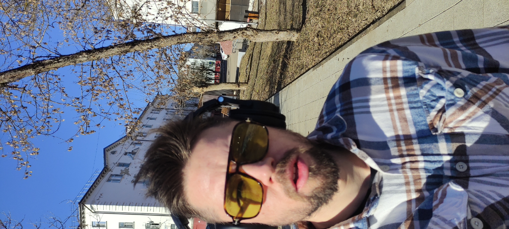 The image is a low-angle, close-up selfie of a man with brown hair and a beard, wearing yellow-tinted sunglasses and a blue, brown, and white plaid shirt. He appears to be wearing headphones. The background includes a bare tree with many small seed pods, a blue sky, a multi-story white building with multiple windows, and a sidewalk bordering a grassy area with dry leaves. The location is likely Fryazino, Russia, based on the prompt. The buildings are likely Soviet-era apartment buildings, and the trees may be linden trees, both typical features of that region. The scene likely depicts a moment outdoors on a sunny day.