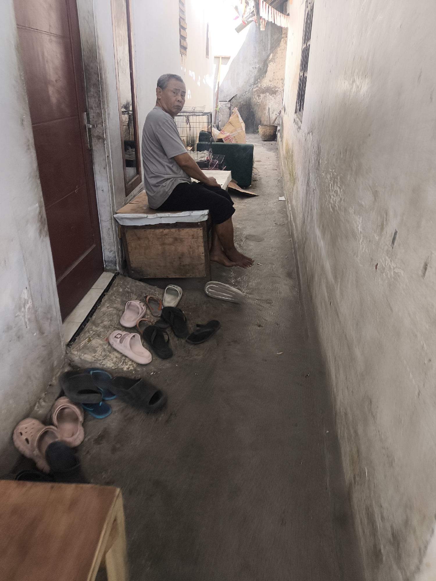 An elderly Asian male sits on a wooden bench in a narrow outdoor alleyway. He is wearing a grey t-shirt and black pants, and his feet are bare. Various pairs of sandals and flip-flops are scattered on the concrete ground in front of him and to his left. To his left is a dark red door. The alley walls are rough and stained. In the background, a woven basket and some cardboard boxes are visible. The scene appears to be in Lumajang, Indonesia.