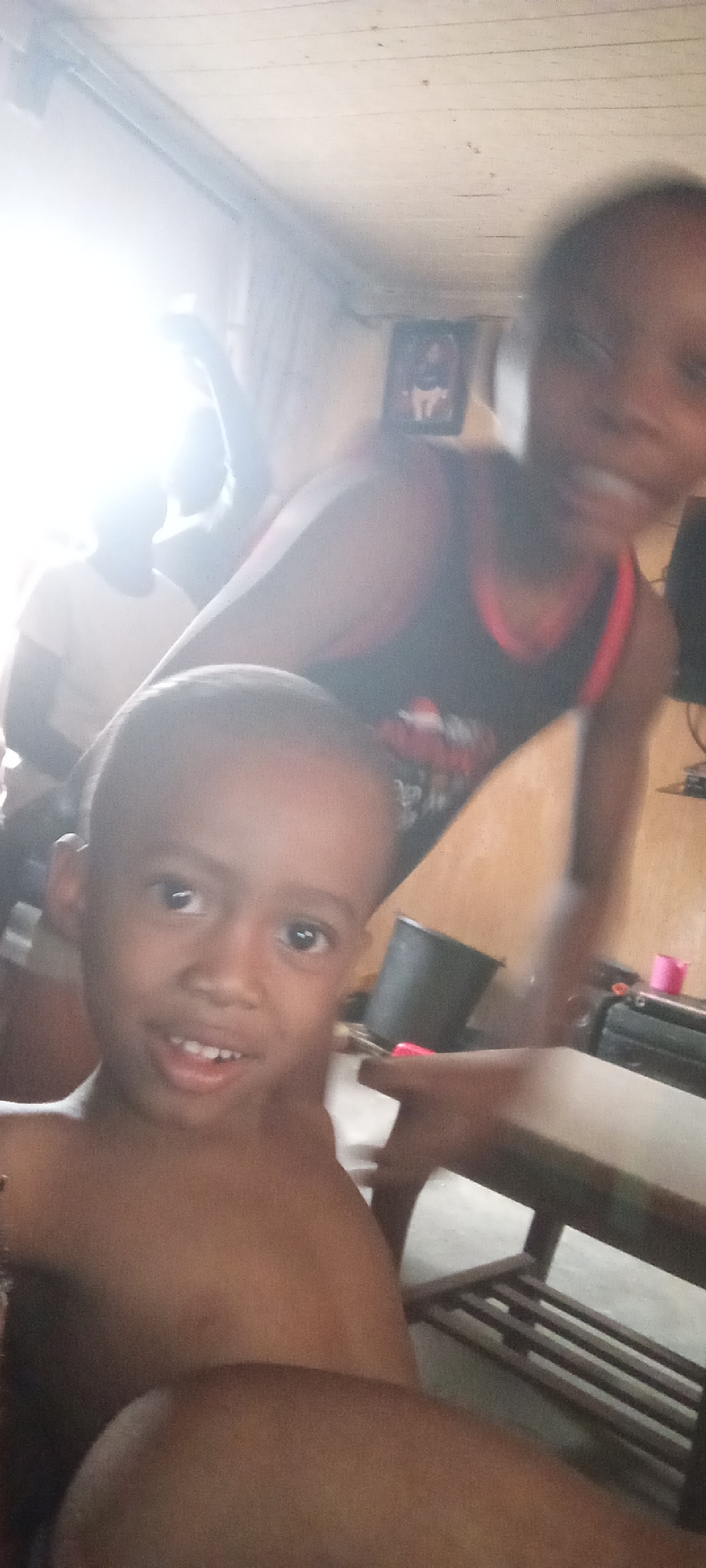 The image shows a young boy in the foreground. Behind him, another child is partially visible, wearing a black tank top with red trim. Additional people are visible in the background near a bright window. The room appears to be indoors, with wooden walls and ceiling. A table is situated to the right of the foreground boy. The location is Unknown, Unknown.
