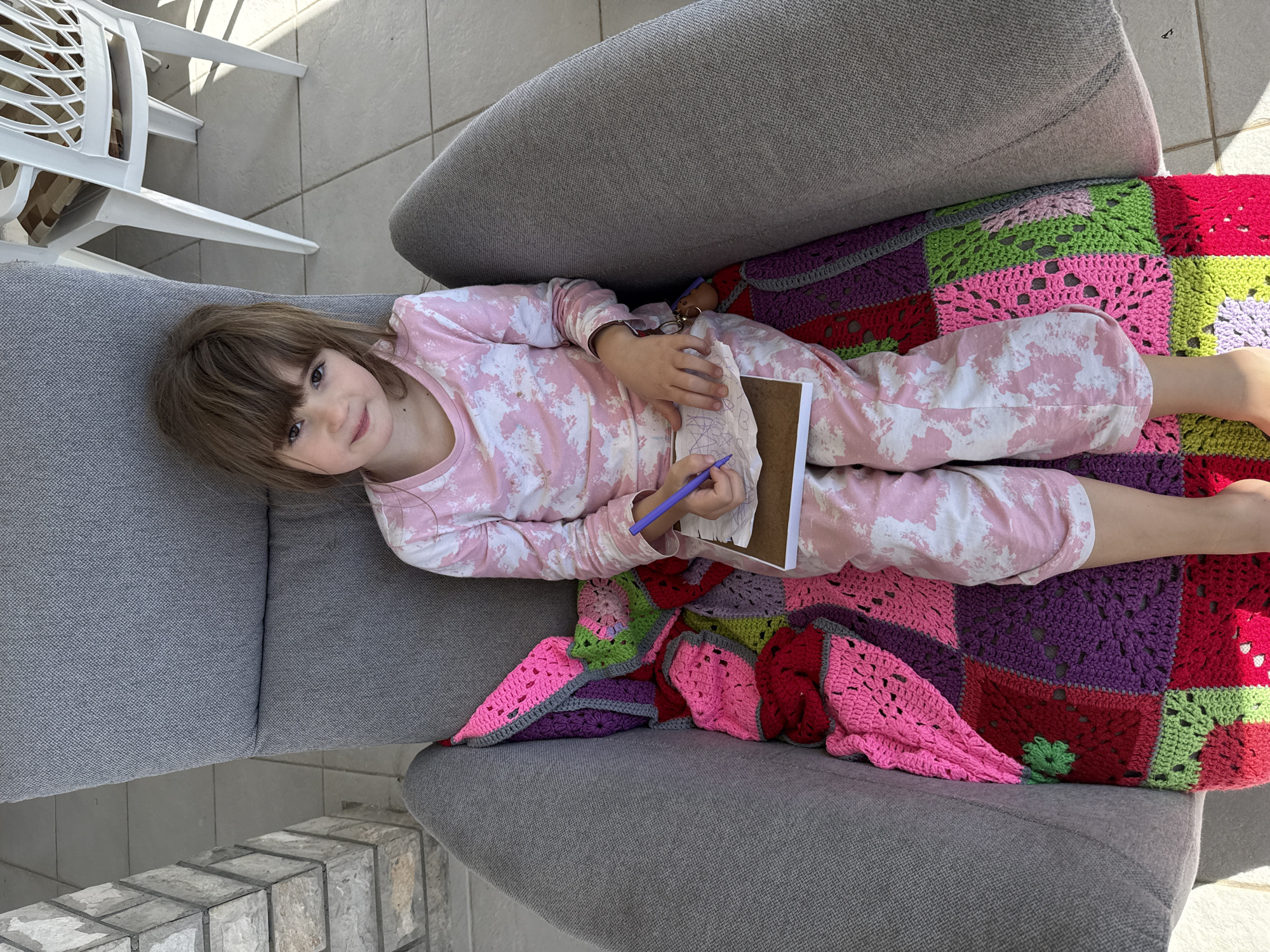 A young girl wearing pink and white pajamas is lying on a gray sofa outdoors, drawing with a blue pen in a notepad. The sofa is covered with a colorful crocheted blanket of pink, green, purple, and red squares. A white plastic chair is partially visible in the upper left corner. The location is likely a patio in Bijela, Montenegro, indicated by the presence of exterior tile flooring and a partial view of an exterior wall.