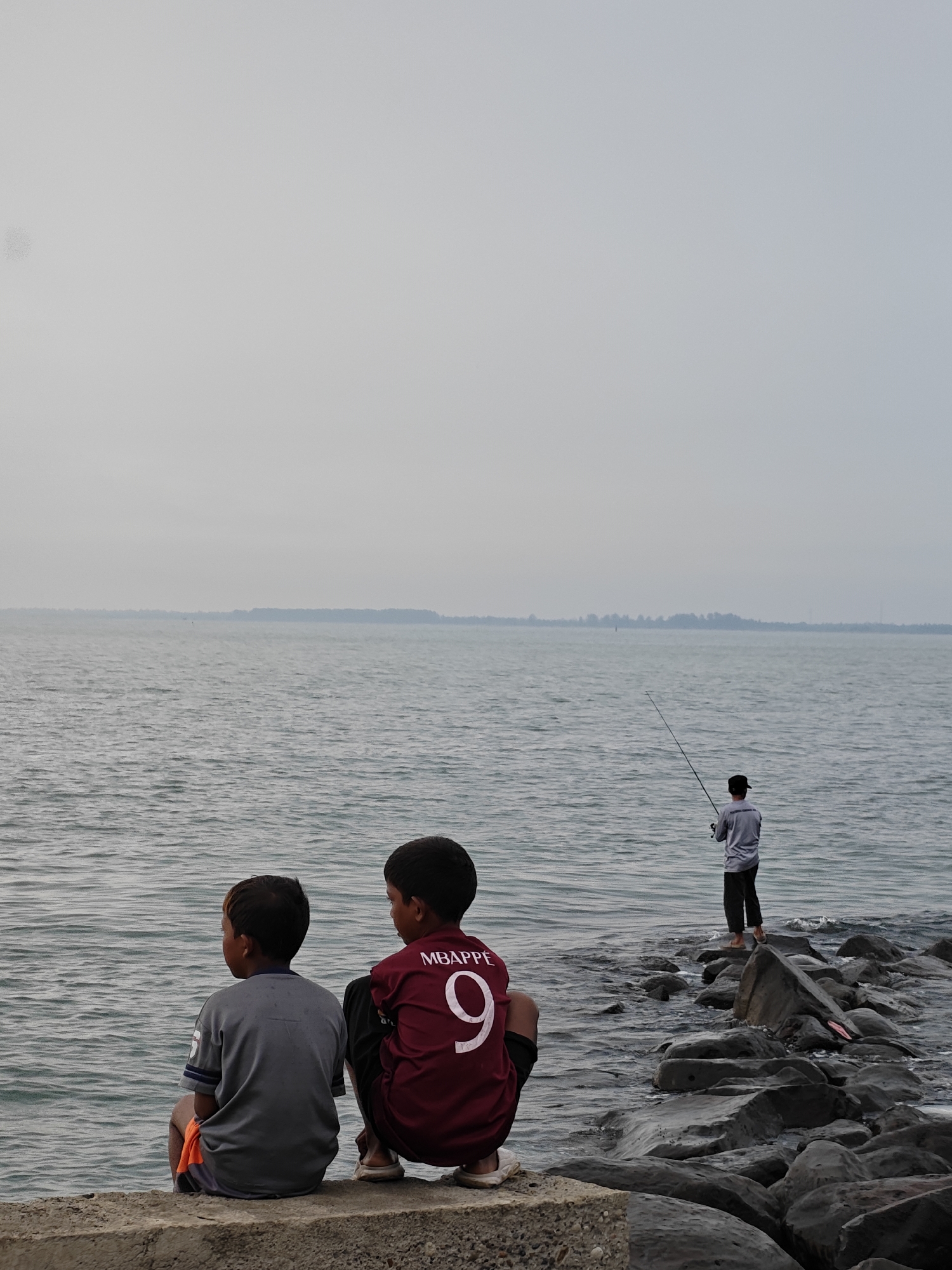 The image depicts an overcast day at the waterside in Lhokseumawe, Indonesia. In the foreground, two young boys are sitting on a concrete ledge, facing away from the camera and looking out at the water. The boy on the left wears a gray t-shirt and orange shorts. The boy on the right wears a maroon t-shirt with the number "9" and the name "MBAPPE" on the back.
Further out into the water, a lone figure stands on a rocky outcrop, fishing with a rod. The sea is choppy, and a distant, tree-lined shore is visible on the horizon. The overall atmosphere is calm and somewhat melancholic, perhaps due to the cloudy weather. The scene suggests a moment of quiet observation and leisure by the sea.