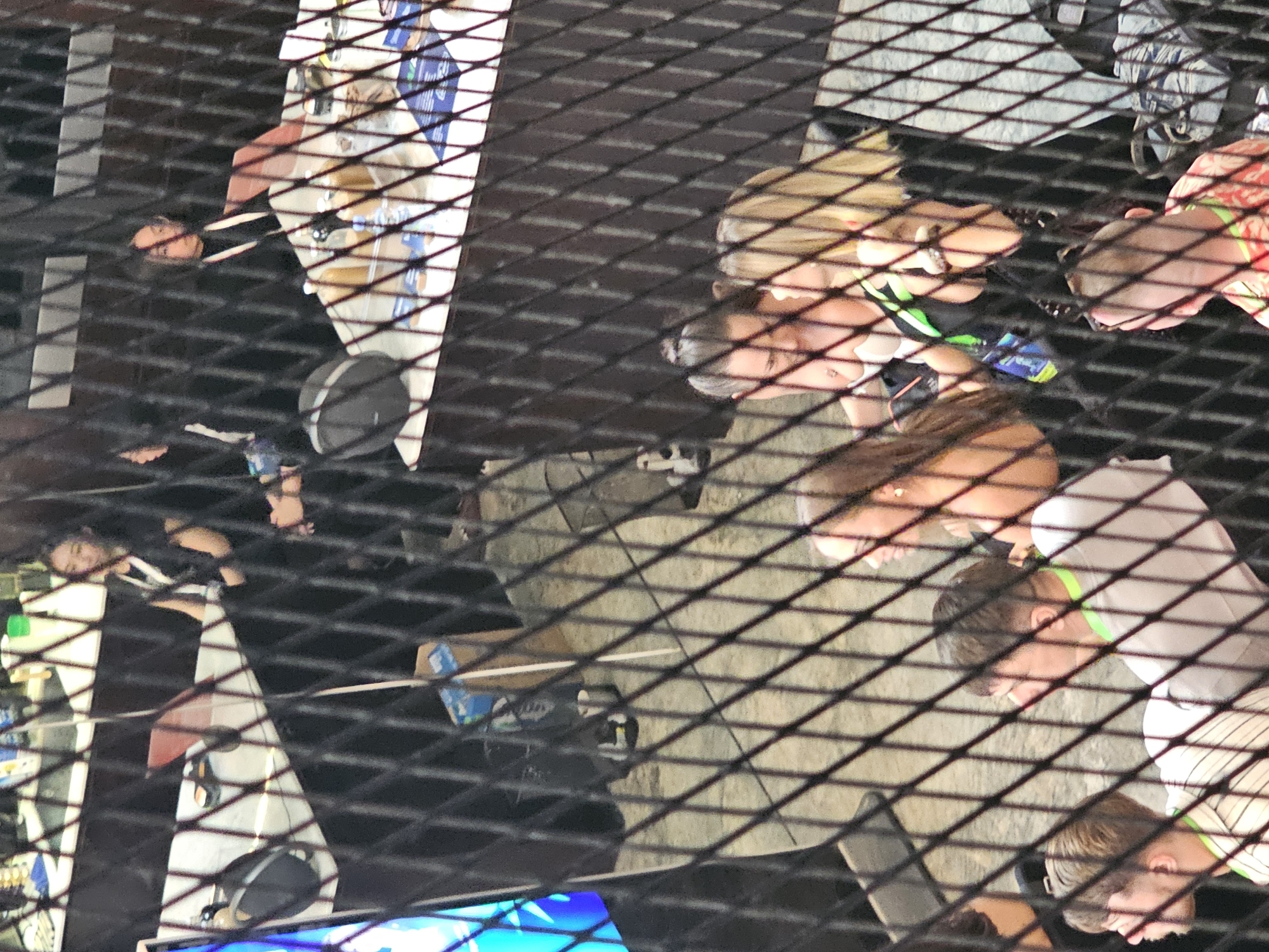 The image, taken from above, shows a group of people gathered around a structure in an outdoor setting. The view is partially obstructed by a metal grid or mesh, creating a patterned overlay. Several individuals can be identified; some are looking downward. One person has blonde hair, and another is wearing a brightly colored wristband. A variety of objects are visible, including what appears to be beverages, containers, and other unidentified items resting on a surface. In the lower part of the frame, there's a glimpse of a brightly lit blue element. Based on the location information (North Kuta, Indonesia), it is likely an outdoor entertainment or dining venue.