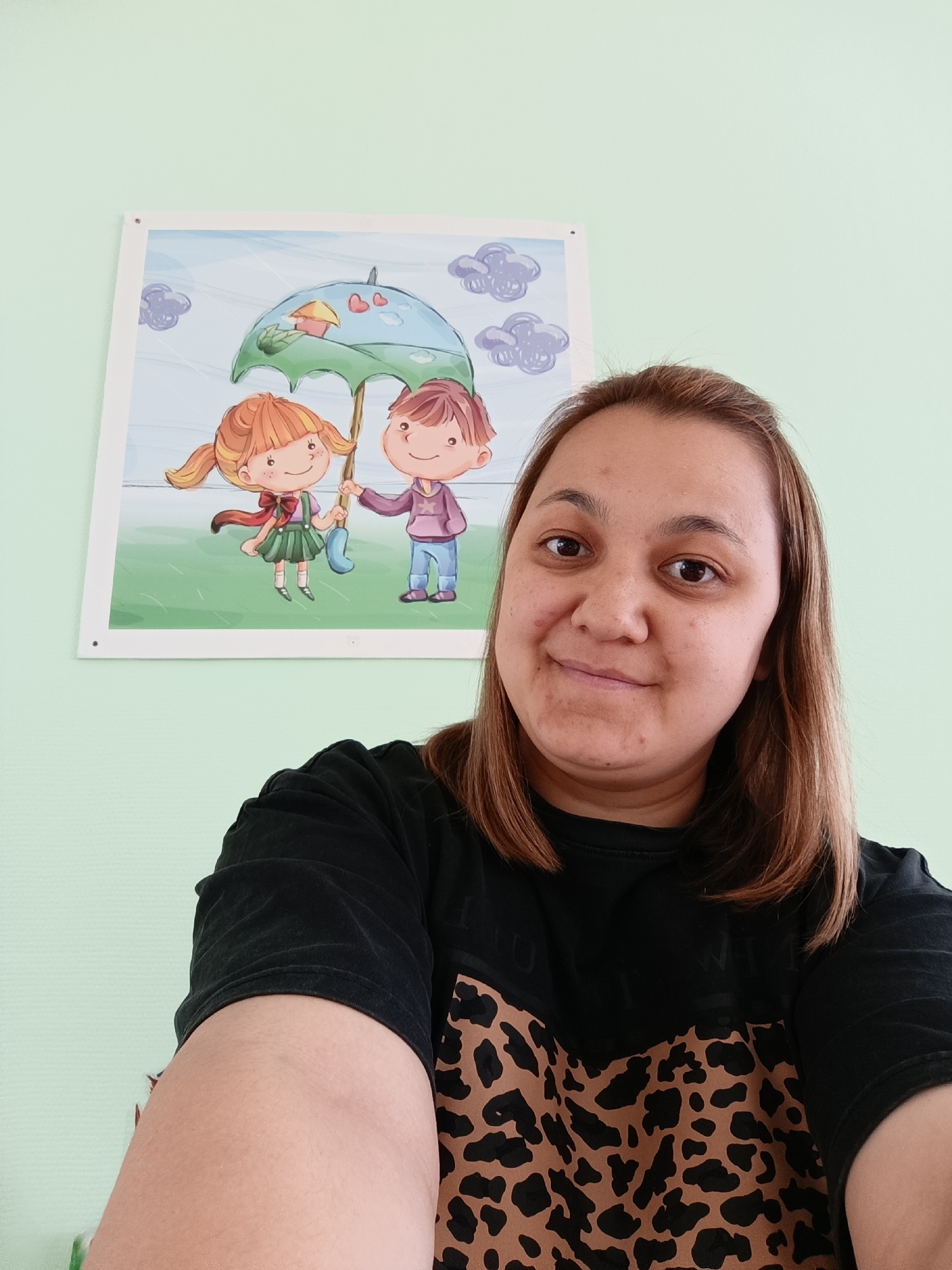 A woman is captured in a selfie indoors, with Chaikovsky, Russia as the contextual location. She has light brown hair and a gentle, slight smile as she looks towards the camera. She is wearing a black t-shirt with a prominent brown and black leopard print pattern across the chest and stomach area.
Behind her is a light green wall. A rectangular picture or poster is affixed to the wall, depicting two cartoon children. The girl, on the left, has reddish-brown pigtails, a green dress, and a red bow. The boy, on the right, has brown hair and wears a purple hoodie and blue pants. Both children are smiling and jointly holding a large green umbrella, which features a small house and red heart shapes on its surface. Grey, stylized clouds are visible in the light blue background of the artwork. The scene appears to be well-lit, suggesting daytime. There is no visible text on the woman's clothing or within the poster's illustration.