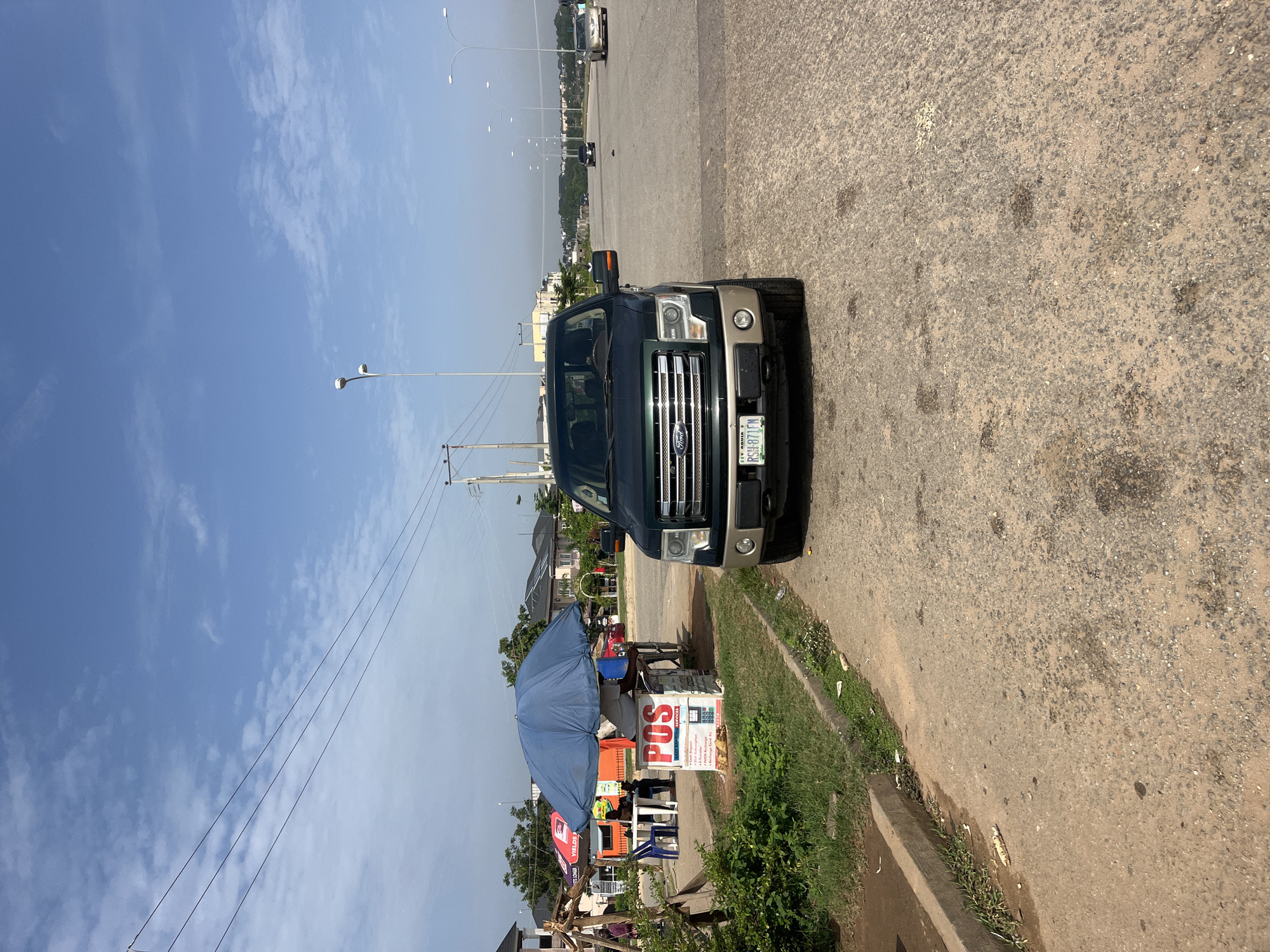 In Karum, Nigeria, a dark green Ford F-150 truck is parked on the right side of a paved road, facing forward. The truck's license plate reads "RSH 871FN". To the left of the truck, a blue umbrella shades a small stand labeled "POS" with various financial services listed. A person is seated behind the POS stand. Behind the stand, there are two orange tents and some tables and chairs. The road itself is wide with multiple lanes and illuminated by streetlights. Further down the road, other vehicles are visible, and residential buildings line the background. The sky is clear with scattered clouds, and the environment appears to be a commercial or roadside area with some greenery on the left.