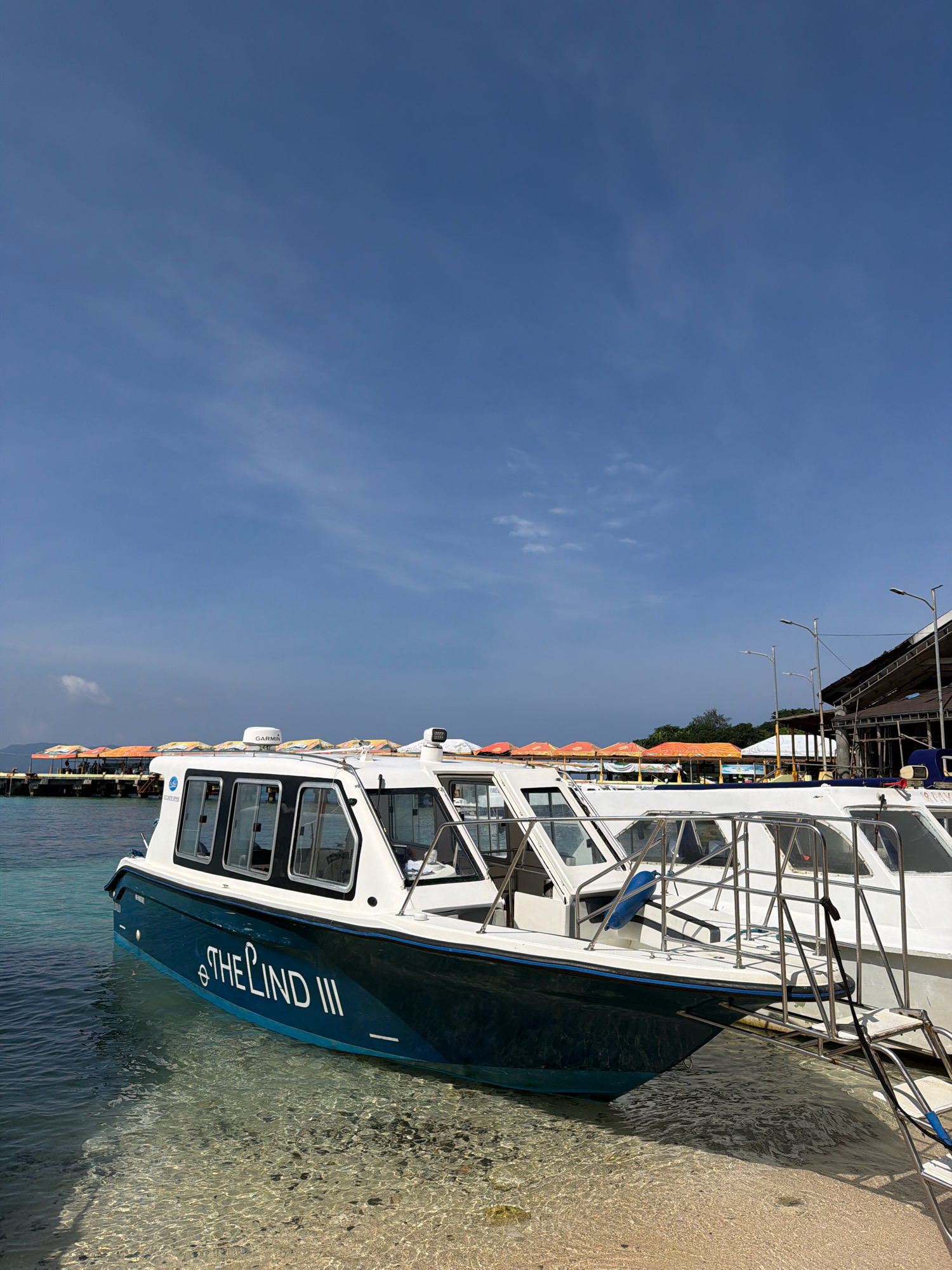 A clear blue sky with wispy clouds stretches above a tranquil body of water. In the foreground, a dark blue and white boat named "THE LIND III" is docked near a sandy shore. The boat features large windows and a metal railing. To the left of the boat, the clear water reveals the sandy seabed. In the background, a pier extends into the water, lined with colorful umbrellas providing shade over seating areas. A building with a dark roof and light poles is visible on the right side of the pier. The scene appears to be at a beach or a port in Nabas, Philippines, a municipality known for its coastal areas and tourist attractions.