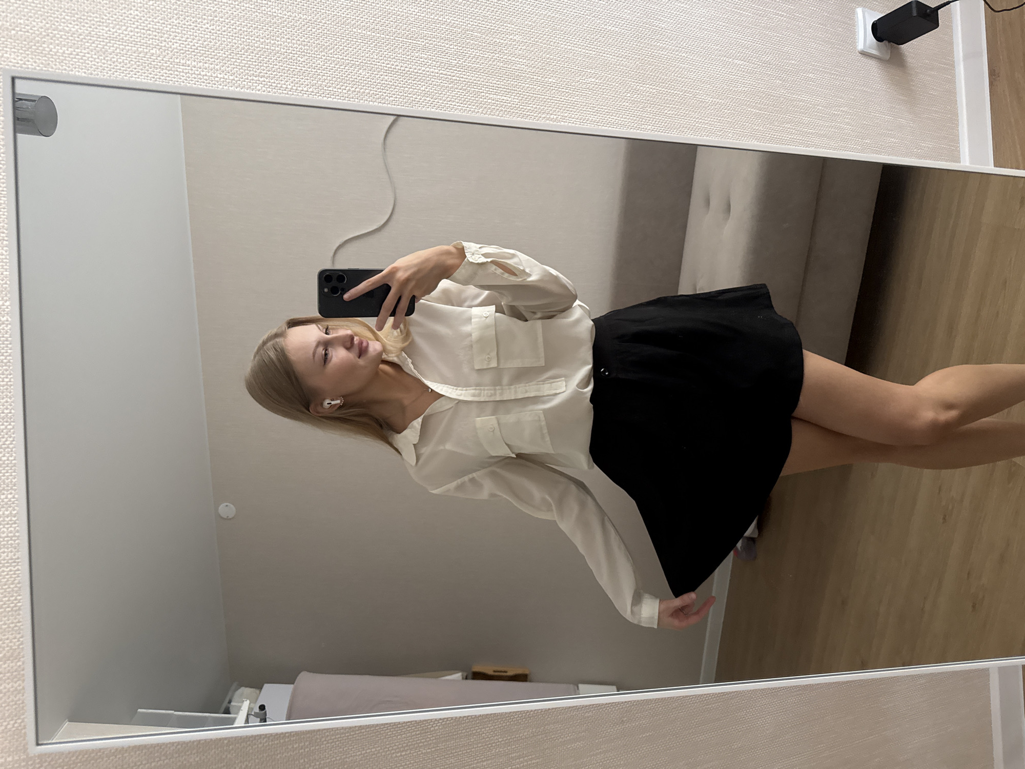 The image is a full-length mirror selfie of a young woman with blonde hair, wearing a white collared shirt and a black skirt. She is holding a black smartphone in her right hand and is taking the picture. The woman is standing in what appears to be a bedroom or dressing area with light-colored walls and wooden flooring. A couch is visible in the background reflection. A white electrical outlet with a black adapter is mounted on the wall to the right. There are no discernible landmarks or indicators to suggest the location is specifically in the city of Perm, Russia.