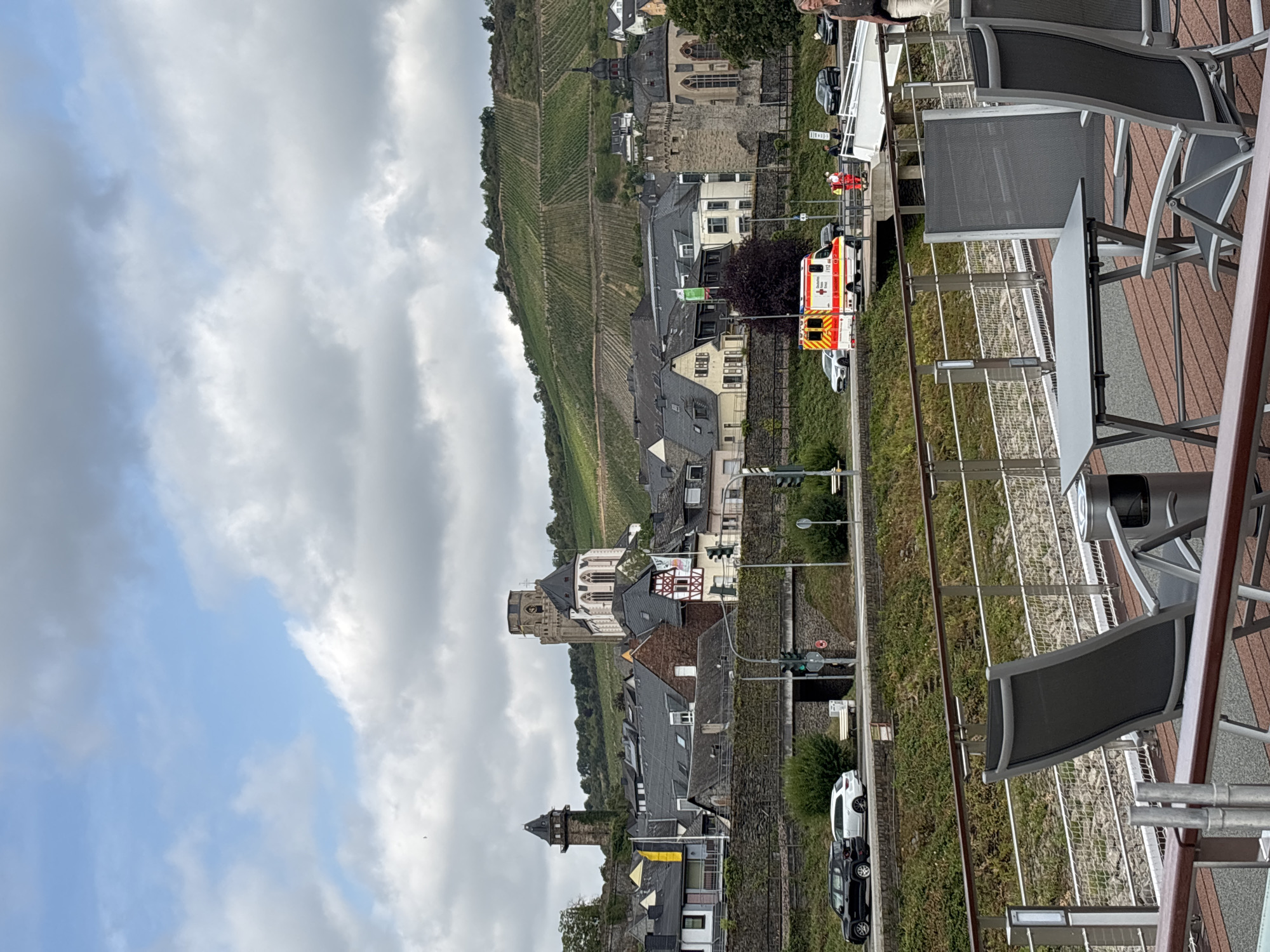 The image captures a view of the town of Oberwesel, Germany, with a focus on its historical architecture and the surrounding landscape. In the foreground, a section of a riverboat's deck is visible, featuring two empty chairs and a small table. Beyond the deck, a stone wall separates the riverbank from a street level. On the street, two cars are parked: a black SUV and a white compact car. Traffic lights are visible, indicating an active road.
Further into the scene, an ambulance with flashing lights is stopped on the side of the road, with two individuals in red vests attending to it. A bridge extends over a lower section of the embankment.
The town itself is characterized by its densely packed buildings with gray tiled roofs. Prominent structures include a tall, dark tower on the left, resembling a watchtower, and a larger building with a bell tower and Gothic architectural elements, possibly a church or cathedral, further to the right. The town is nestled against a steep hillside covered in meticulously maintained vineyards, indicative of the region's wine production. The sky is overcast with scattered clouds. The overall impression is one of a historic European town situated in a scenic, rural setting.