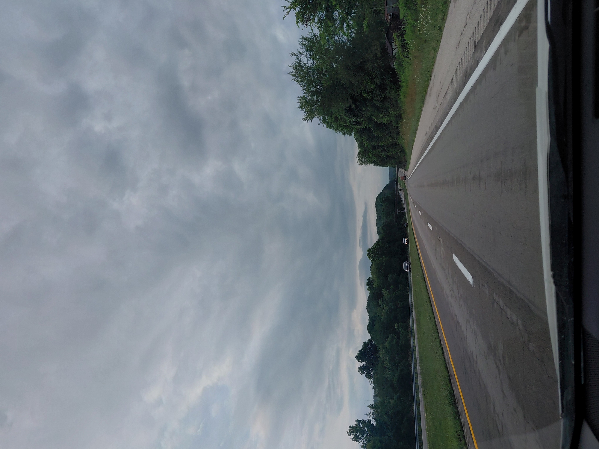 The image captures a view from a moving vehicle on a highway, likely in or near Clay City, United States of America, under a cloudy sky. The highway has multiple lanes with clear white and yellow lane markings. Green trees and foliage line both sides of the road, with some occasional residential structures visible on the right. A few cars are present on the highway, indicating moderate traffic. The overall atmosphere is overcast, suggesting a possibility of rain or an impending storm.
