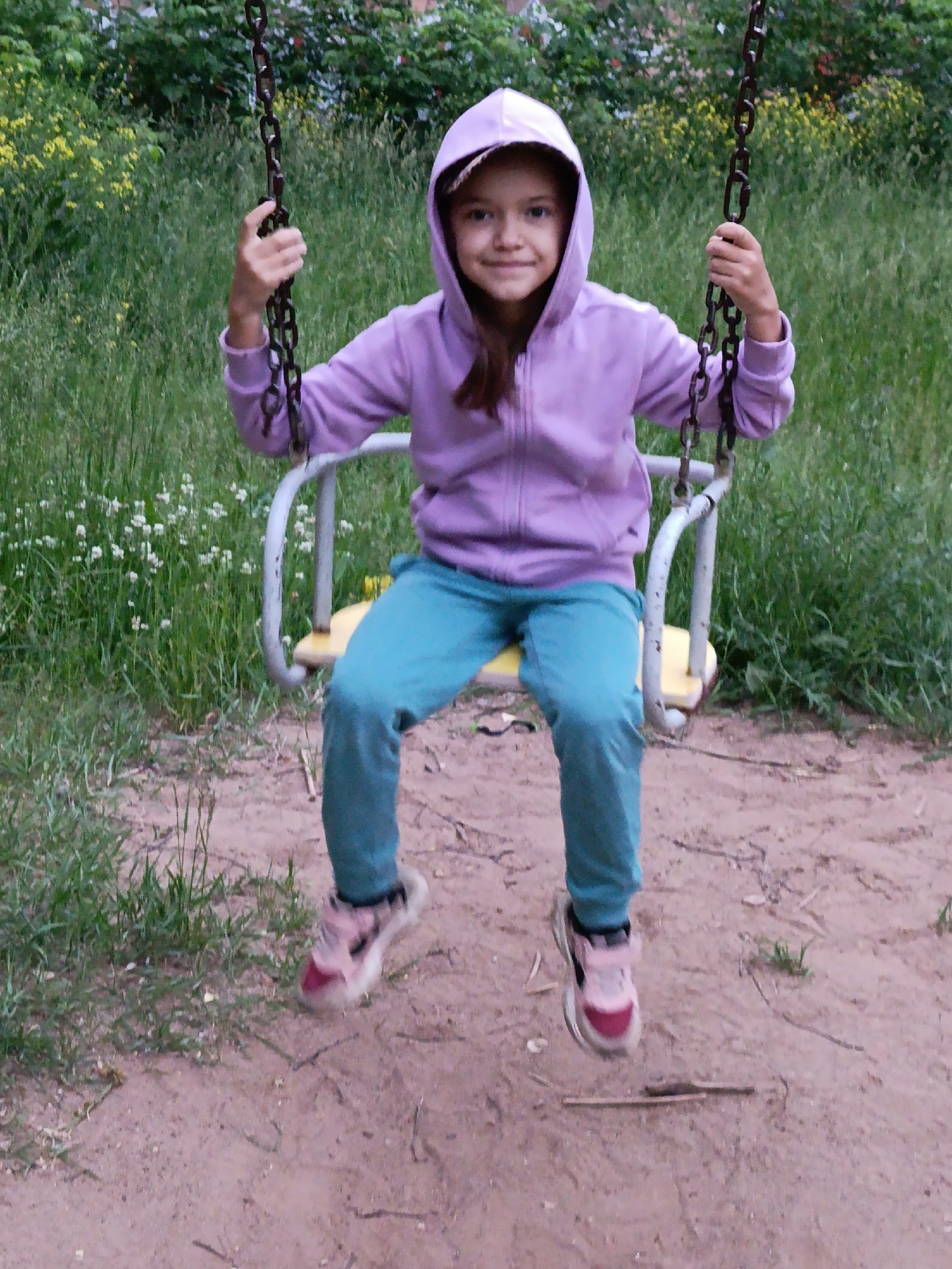 A young girl is sitting on a swing set in a park in Chaikovsky, Russia. The girl is wearing a purple hooded sweatshirt and blue pants. She is holding onto the chains of the swing with both hands and smiling at the camera. The swing set is made of metal and has a yellow seat. In the background, there is green grass, trees, and some yellow flowers. The ground is covered with sand and dirt. The weather appears to be overcast.