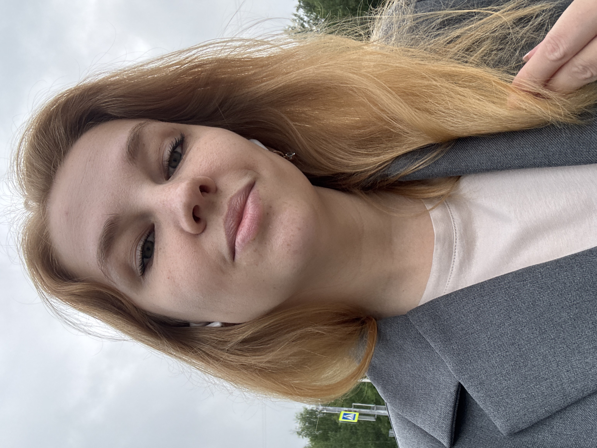 A young woman with shoulder-length, reddish-blonde hair, wearing a grey blazer over a light-colored t-shirt and white earbuds, is captured in a close-up selfie. Her face is angled slightly, and she appears to be outdoors with an overcast sky above. In the background, a green tree line and a yellow pedestrian crossing sign are visible. The setting is indicative of a street scene in the city of Zavolzhye, Russia, likely a suburban or park area given the foliage and pedestrian infrastructure.