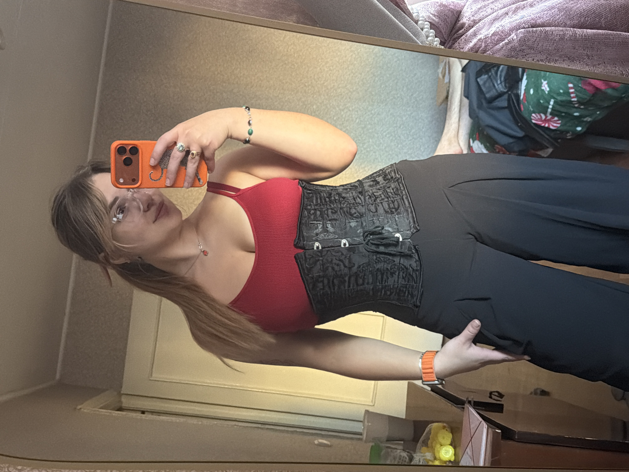 A woman takes a selfie in a mirror indoors. She is wearing glasses, a red top, a black corset-like garment over the top, and black pants. She holds an orange smartphone in her left hand. She has jewelry on her left hand and wrist, and an orange watch on her right wrist. The background includes a tan wall, a white door, and a dresser top with various items. The location of the photo in Naberezhnye Chelny, Russia is not discernible.