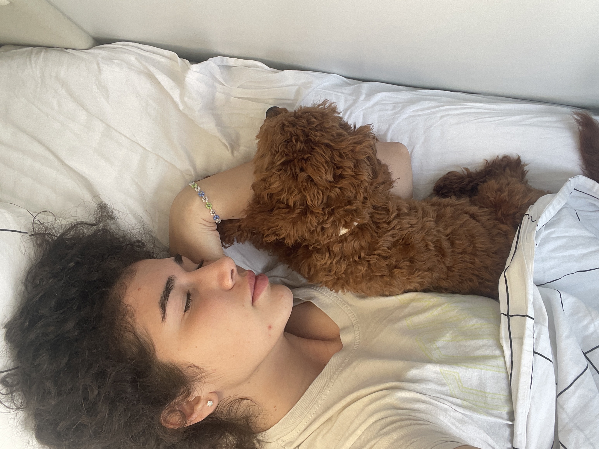 A young person with dark, curly hair and a small, reddish-brown poodle-type dog are depicted sleeping peacefully together on a bed. The person, appearing to be female, is lying on her right side, eyes closed, with her head resting on a white pillow. She wears a light-colored short-sleeved t-shirt with a subtle light green geometric pattern and has a small stud earring in her visible ear and a multi-colored beaded bracelet on her left wrist. The dog, with dense curly fur, is nestled closely against the person's left arm and torso. White bed sheets and a duvet with a black grid pattern cover portions of both subjects. The immediate environment is an indoor bedroom with a plain white background. The image contains no discernible visual markers specific to a location in Koprinka, Bulgaria.