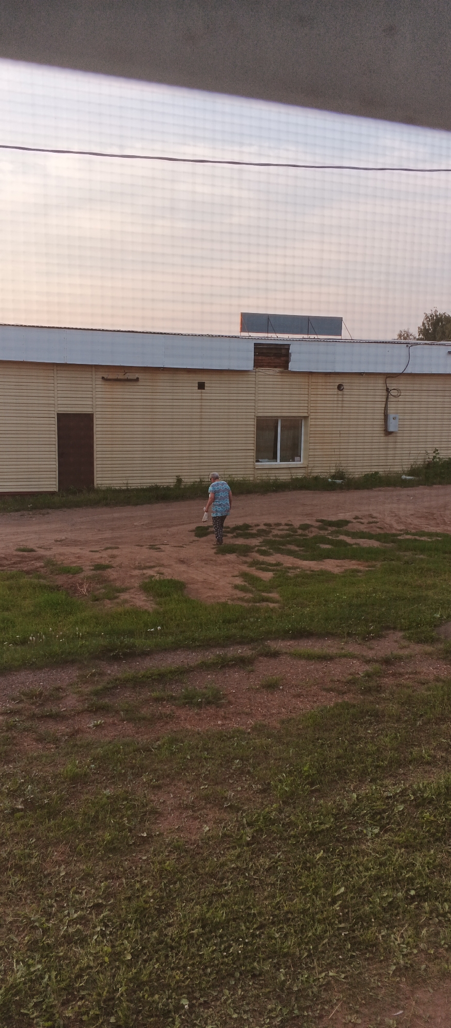 A person wearing a blue and white patterned shirt and dark pants is walking away from the camera in front of a long, light yellow building. The person is carrying a white object. The building has a brown door on the left and a window with white framing on the right. The building's exterior is made of horizontal siding. Above the building, a solar panel is visible on the roof. The ground in front of the building is a mixture of dirt and grass. The sky above is a pale orange and grey. The image appears to have been taken through a screen or window due to the grid pattern visible in the upper portion of the image. The scene is set in an outdoor industrial or commercial area, likely in the city of Podgornoye, Russia.