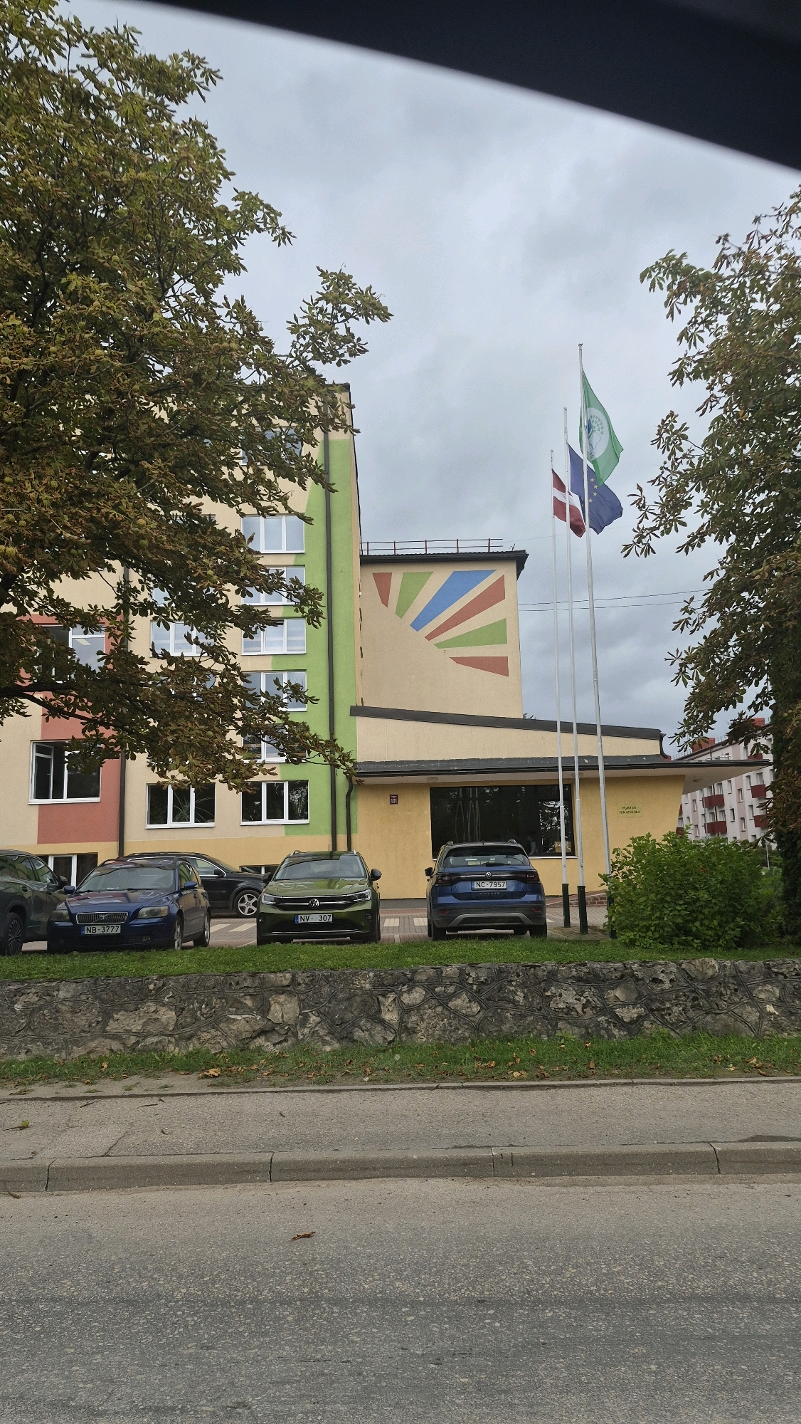 The image shows the front of a building with a colorful mural of a rising sun on its side. Three flags are flying in front of the building: the flag of Latvia, the European Union flag, and a green flag with a white emblem. Several cars are parked in front of the building. The building appears to be a school or public institution, given the presence of flags and a mural. The surrounding environment includes trees, grass, and a road. The sky is overcast, suggesting a cloudy day. Based on the presence of the Latvian flag and the architecture, the location is likely in Pļaviņas, Latvia.