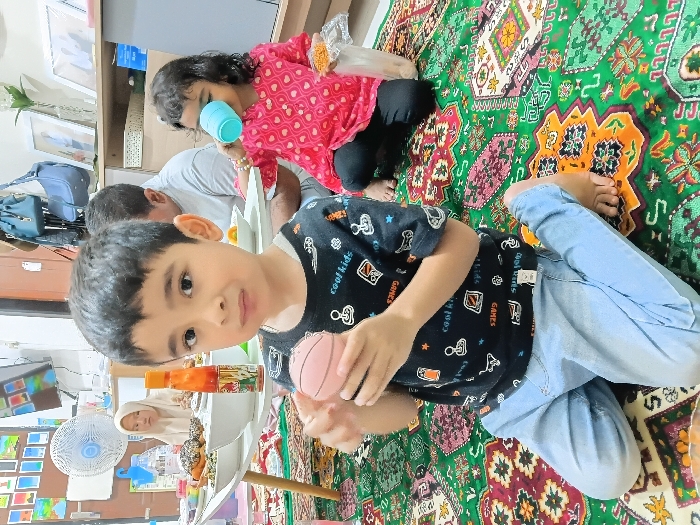 The scene depicts an indoor gathering in Meunasahbaet, Indonesia. A young boy, approximately 3-5 years old, kneels barefoot in the foreground on a vibrant green, red, and orange patterned rug. He wears a black t-shirt with white graphics and light blue jeans, holding a small, light pink sphere (resembling a toy basketball) in both hands, looking directly at the camera with a neutral expression. To his right and slightly behind him, a young girl of similar age sits barefoot on the rug, wearing a pink long-sleeved shirt with white hearts and black pants. She holds a light blue plastic cup to her mouth, appearing to drink, with a clear plastic bag of yellow crispy snacks beside her. In the background, a low white table is laden with bowls of food, including pastries and a red-labeled bottle containing orange liquid. Behind the table, a man in a grey t-shirt is partially visible, and to the left, a woman wearing a beige hijab is also partially visible, both seated on the floor. A white pedestal fan stands to the left of the table. The background includes a wall-mounted television, assorted framed pictures, and hanging bags, indicating a residential setting.