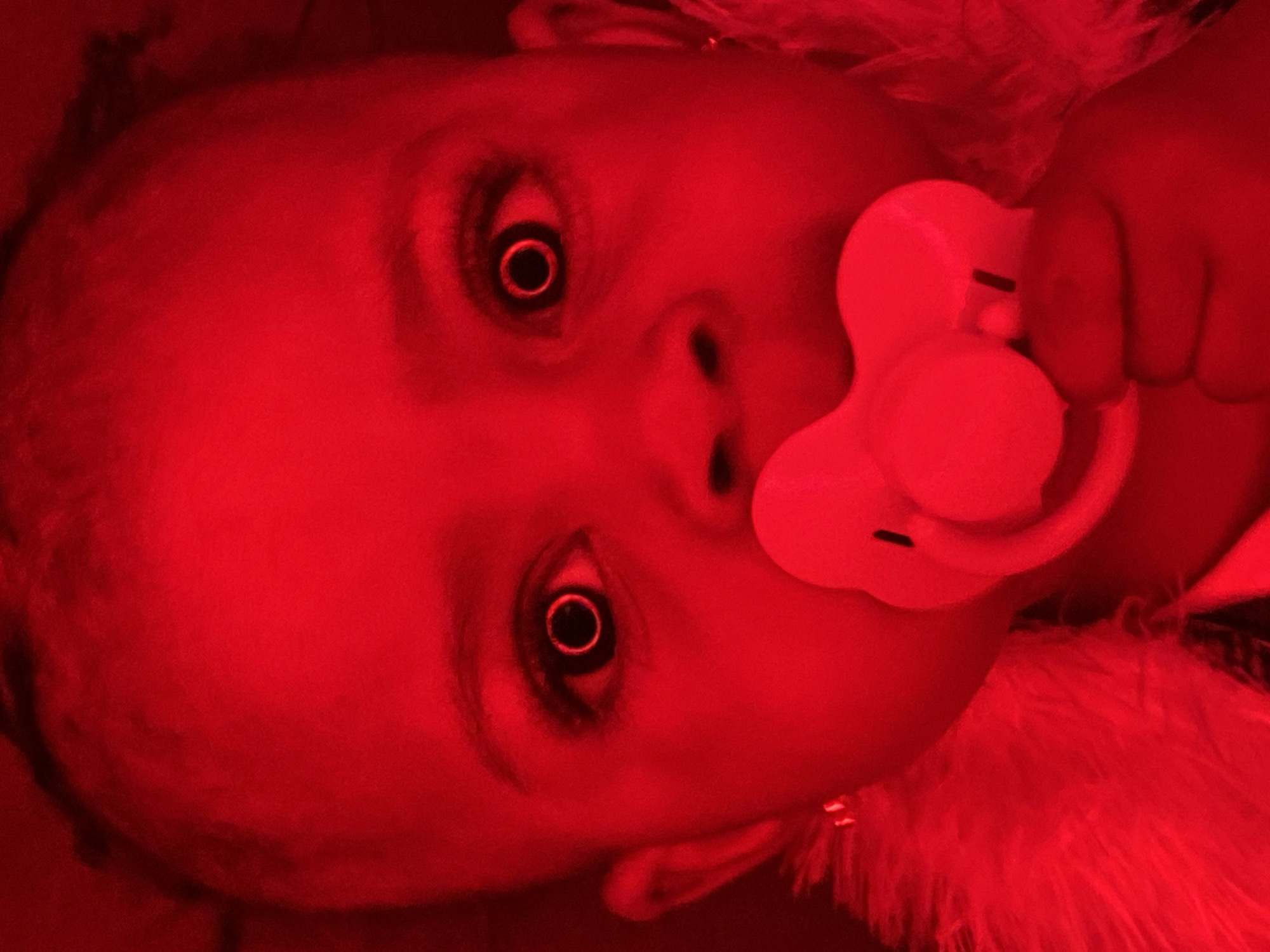 The image is a close-up, red-tinted photograph of a baby's face. The baby has dark skin and is looking directly at the camera with wide eyes. A light-colored pacifier is in the baby's mouth. The baby is wearing a fluffy white garment around its neck, possibly a boa or a collar. The background is dark and out of focus. There is no discernible information within the image to indicate a specific location in Abakaliki, Nigeria.